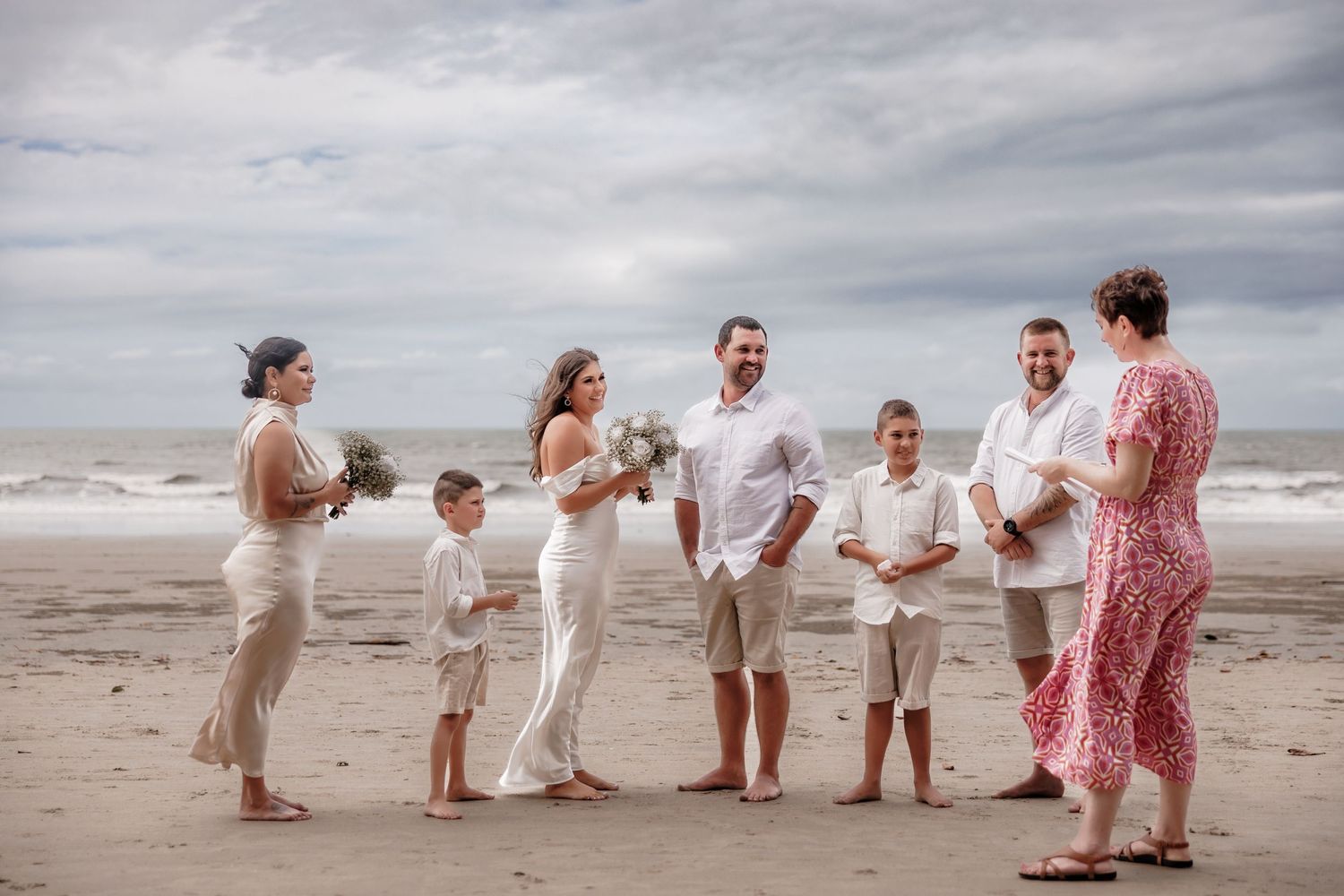 Family gathering on a beach for casual wedding ceremony under cloudy skies.