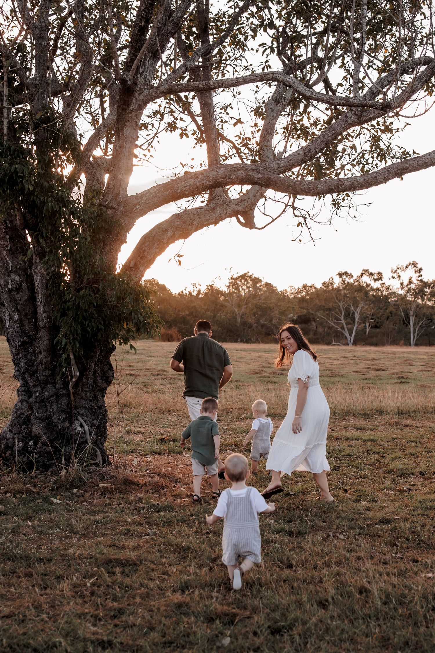 A family walks together under a large tree at sunset in a rustic field during golden hour.