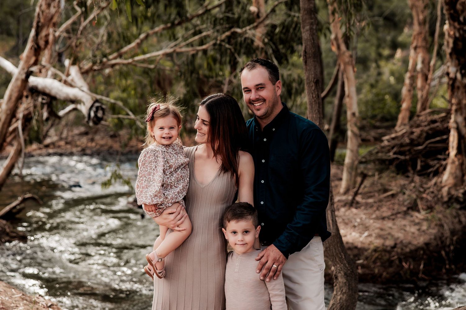 A family poses together by a rocky stream in a wooded setting during sunset.