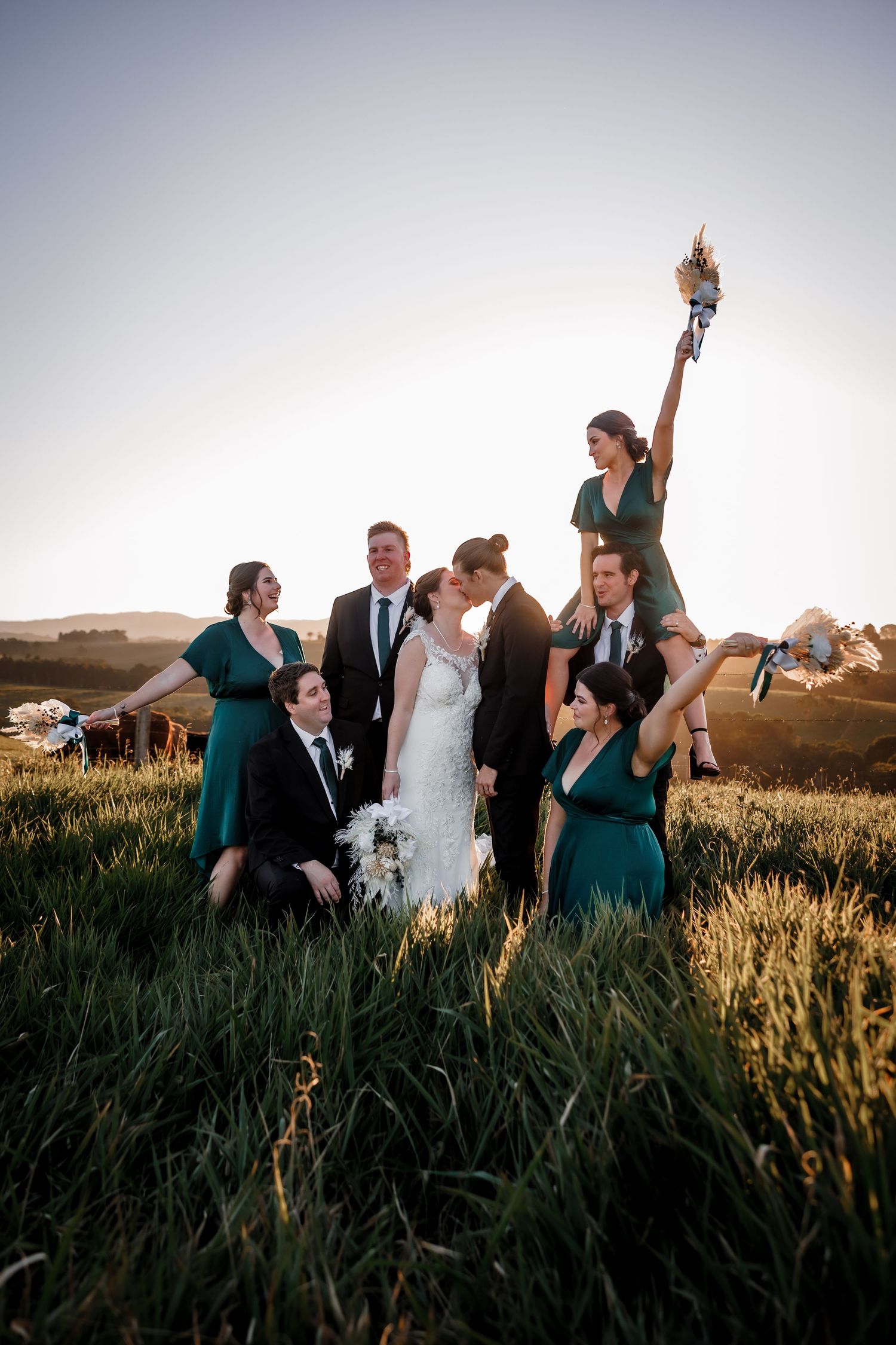 Playful wedding party posing in a grassy field at sunset with dramatic lighting.