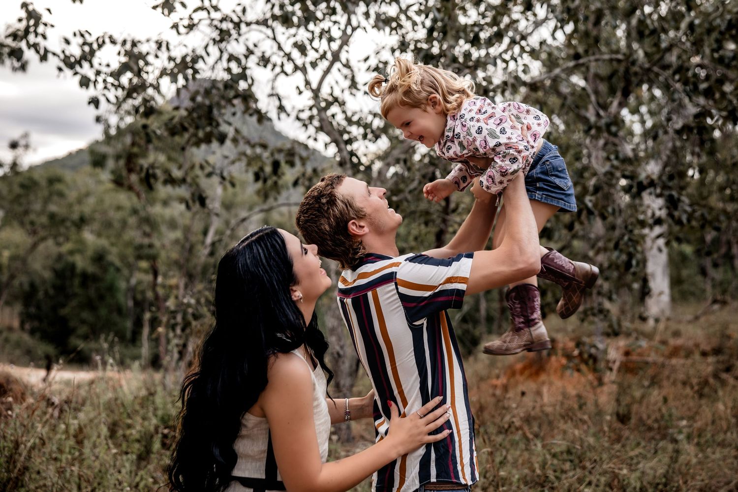 A couple lifts their young child together in a sunny outdoor setting surrounded by trees and natural scenery.