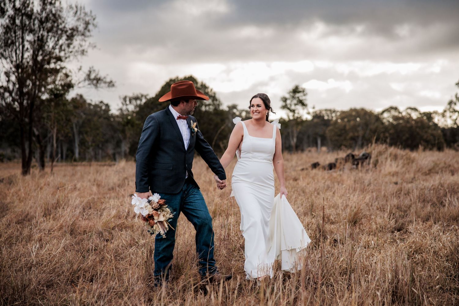 A newlywed couple walks hand in hand through a rustic field at sunset.