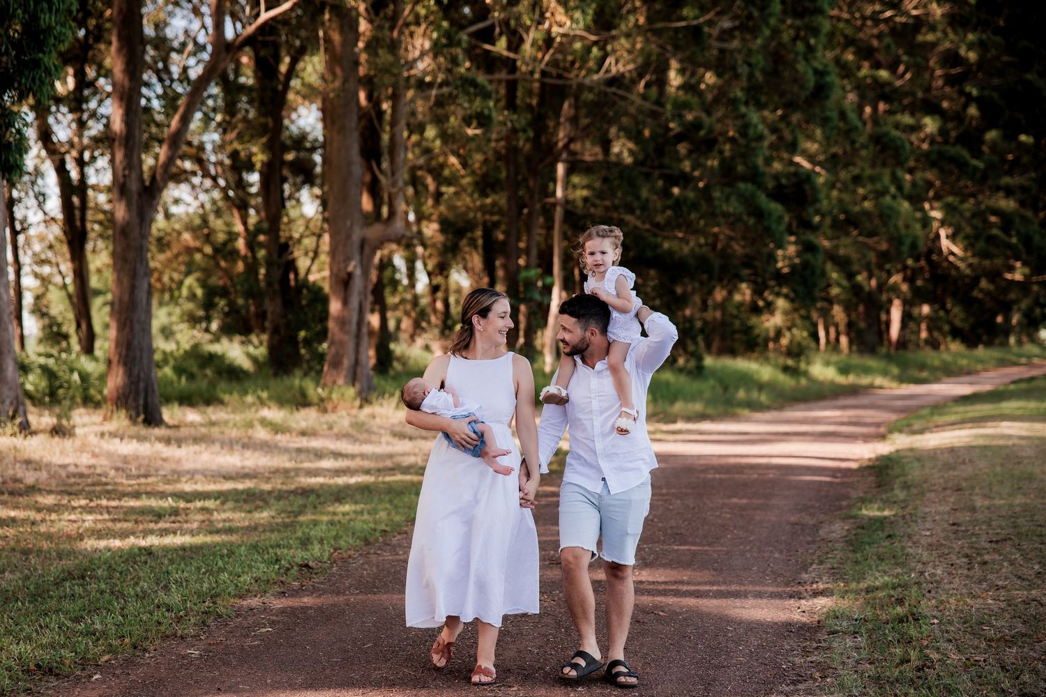 A family walks together on a sunlit forest path, wearing white summer outfits and enjoying a peaceful moment in nature.