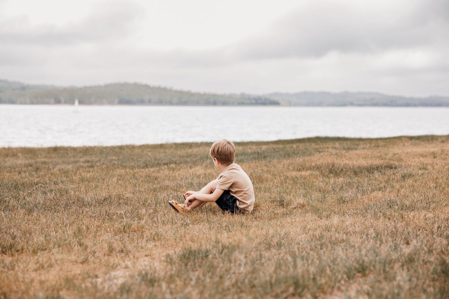 Solitary figure sitting in a grassy field overlooking a serene lake and mountains.