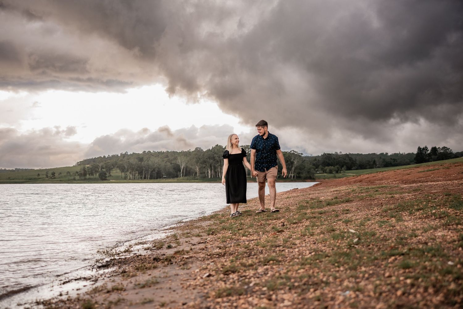 Couple standing on shoreline against dramatic stormy sky and mountain backdrop.