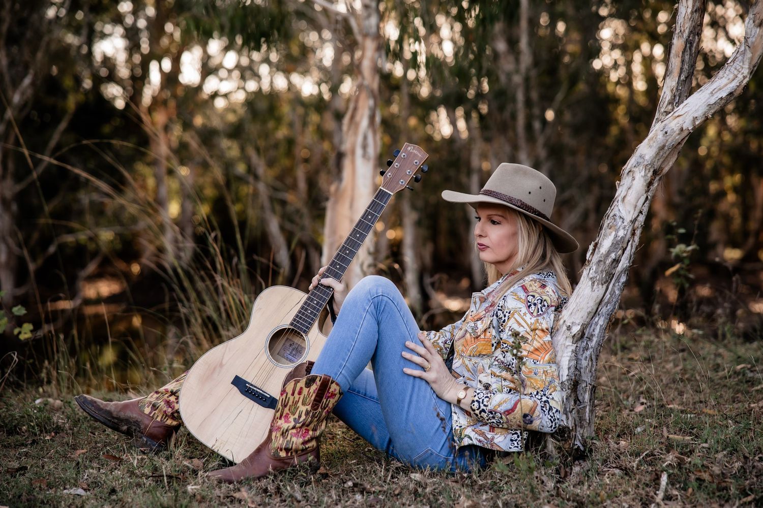 Someone wearing jeans and a floral blouse relaxes against a tree with an acoustic guitar in a woodland setting.