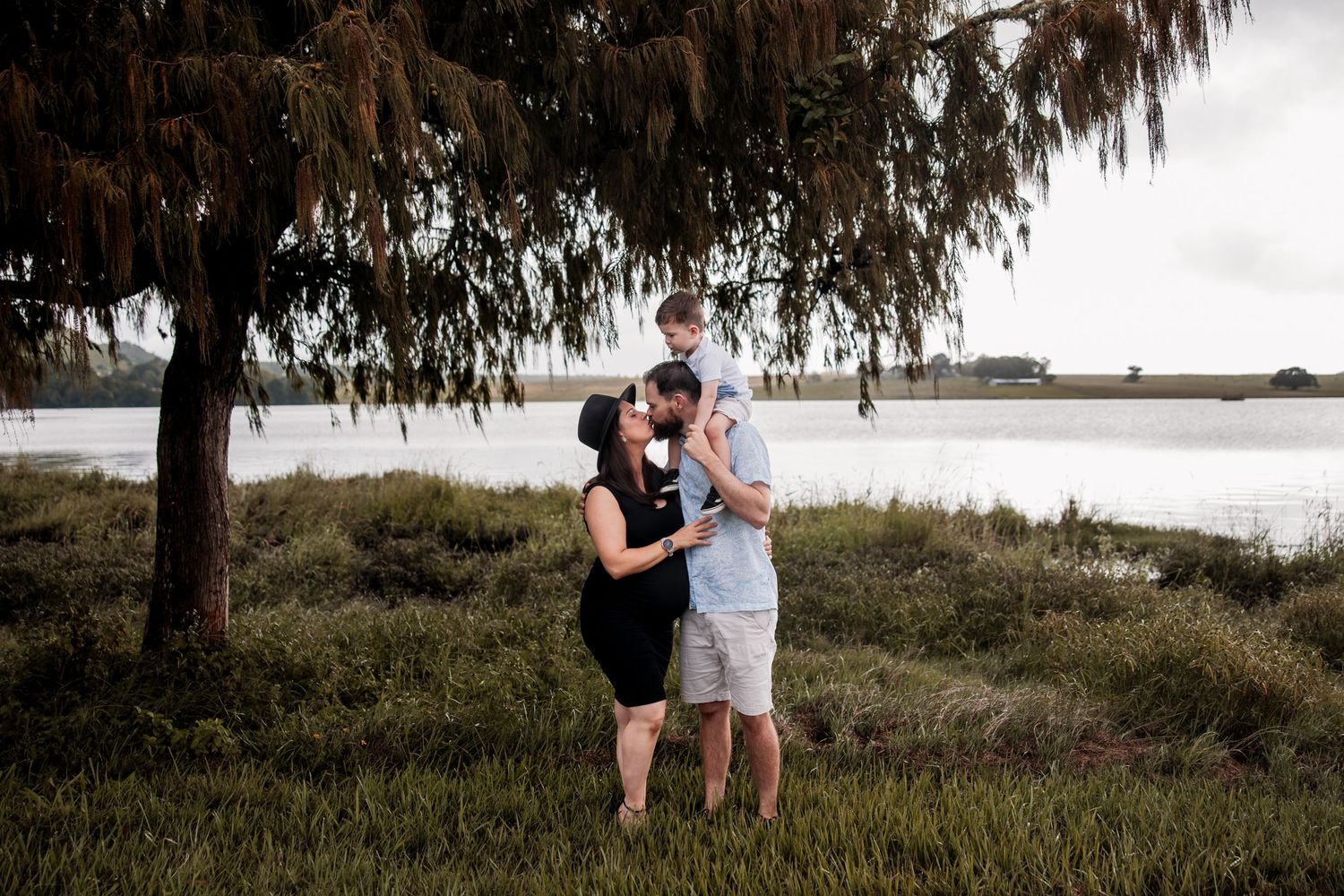 Couple sharing intimate moment under tree branches near waterfront.