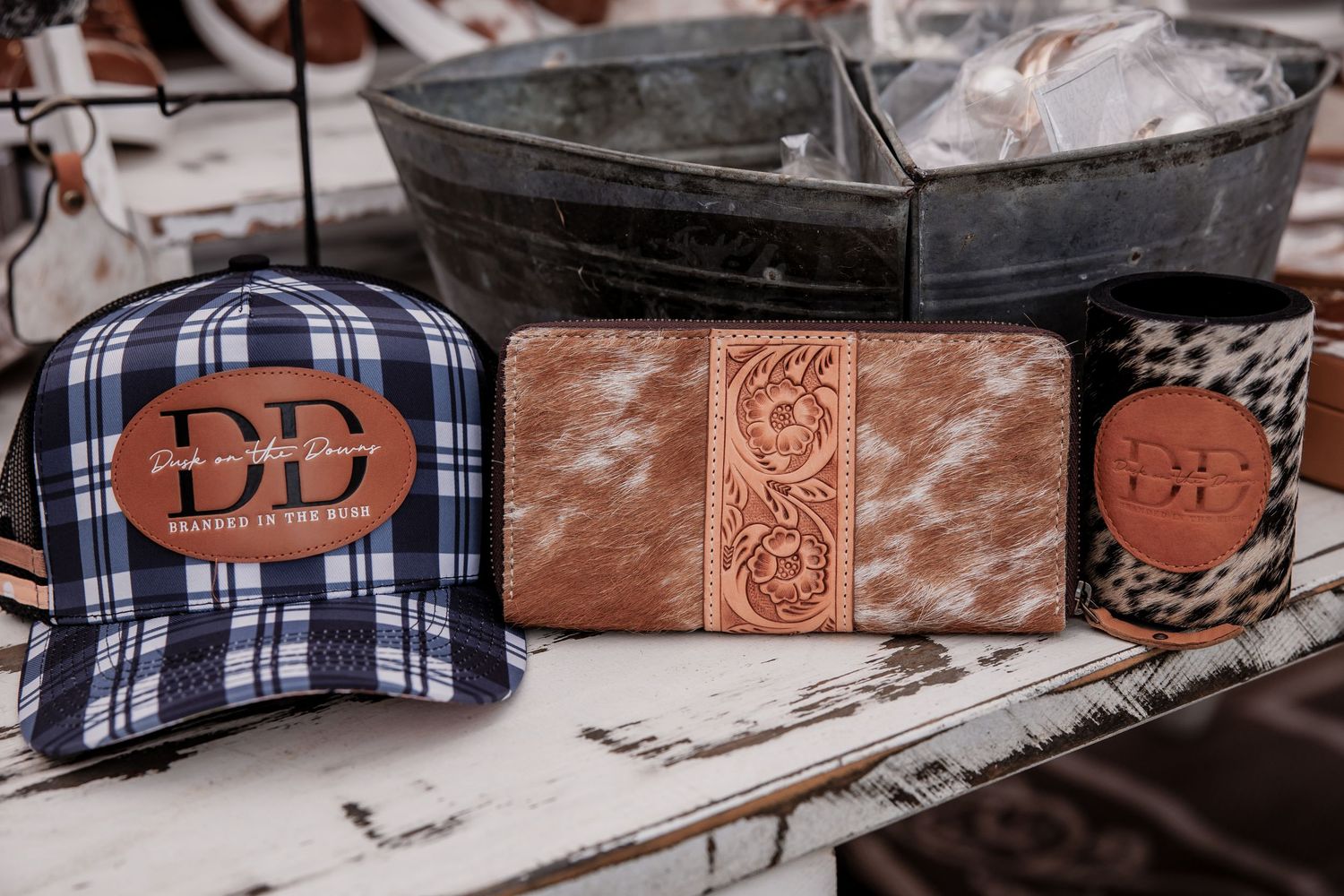 Rustic western accessories including blue plaid cap, brown leather wallet, and cowhide patch on wooden surface.