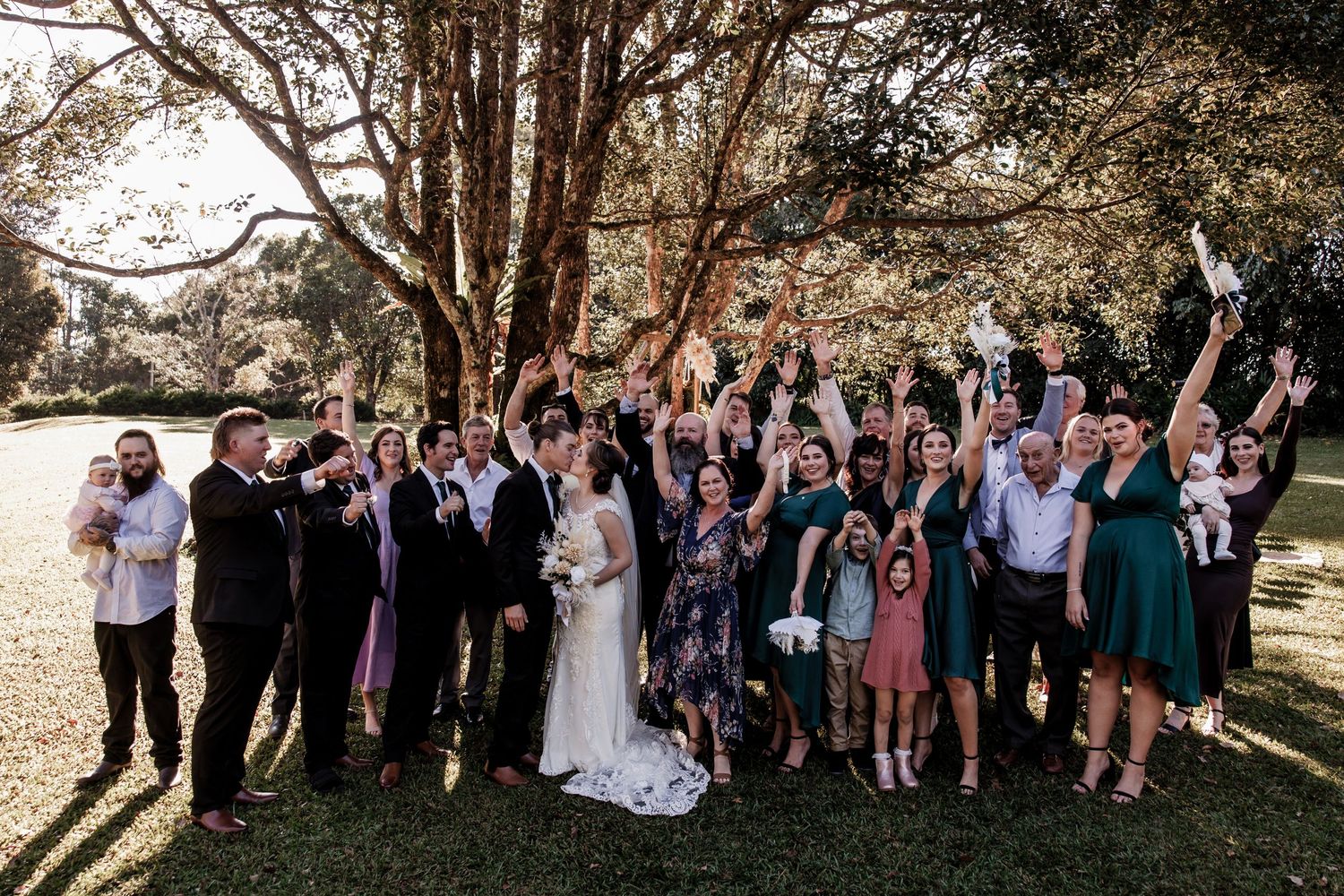 Large wedding party celebrating with arms raised on a sunny lawn surrounded by trees.