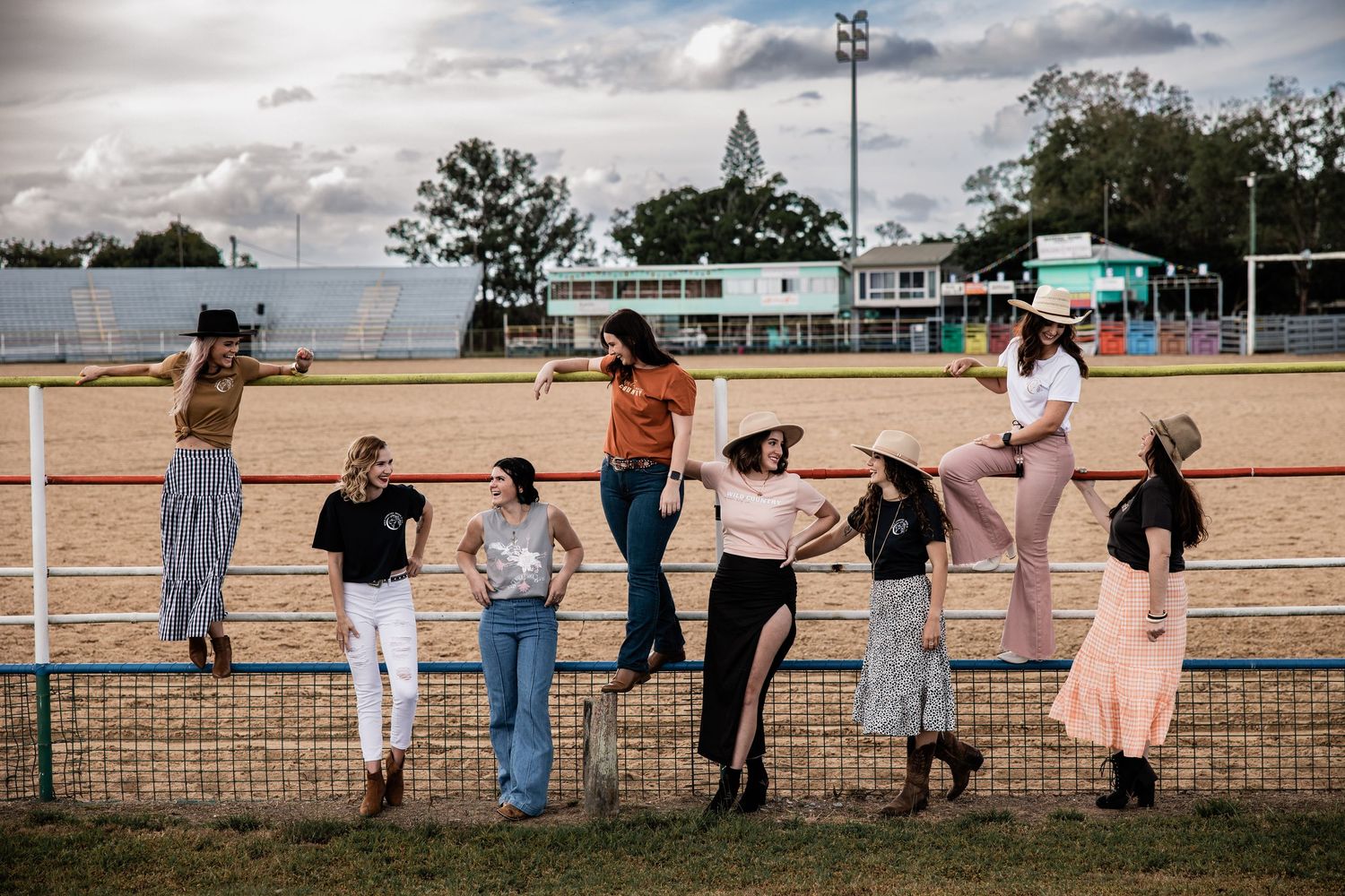 Group of people leaning on a metal fence at a dusty fairground with stadium lights and buildings in the background.