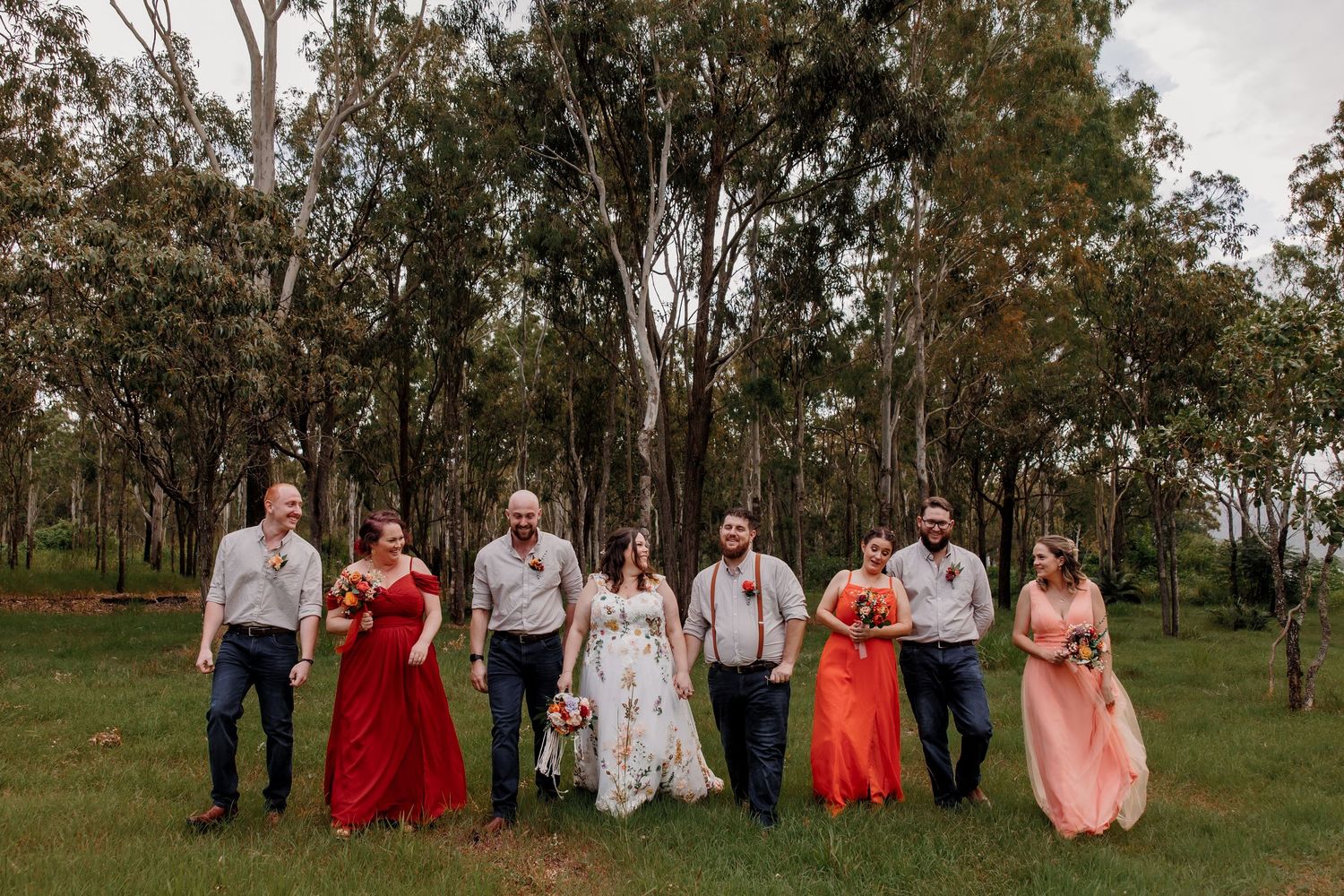 A wedding party in red and pink dresses poses together on grass with trees in the background.