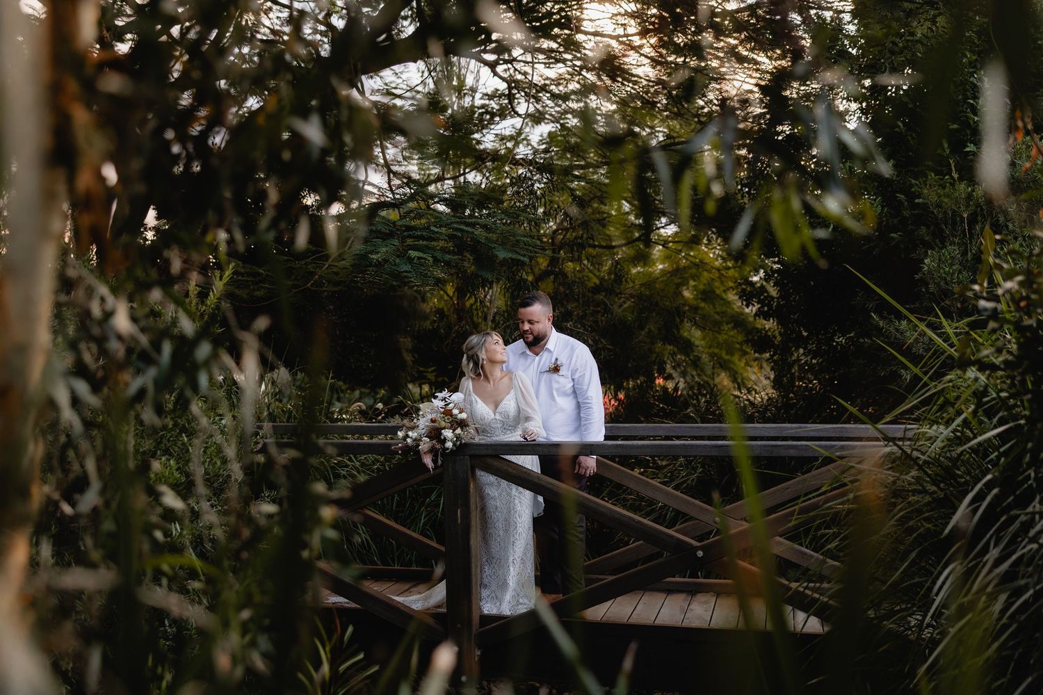 A couple in wedding attire stands on a wooden bridge surrounded by lush tropical greenery.