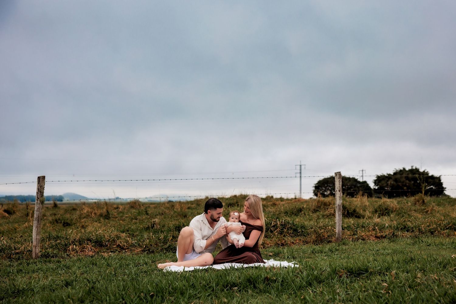 Two people share an intimate moment while picnicking on a blanket in a grassy coastal field under cloudy skies.