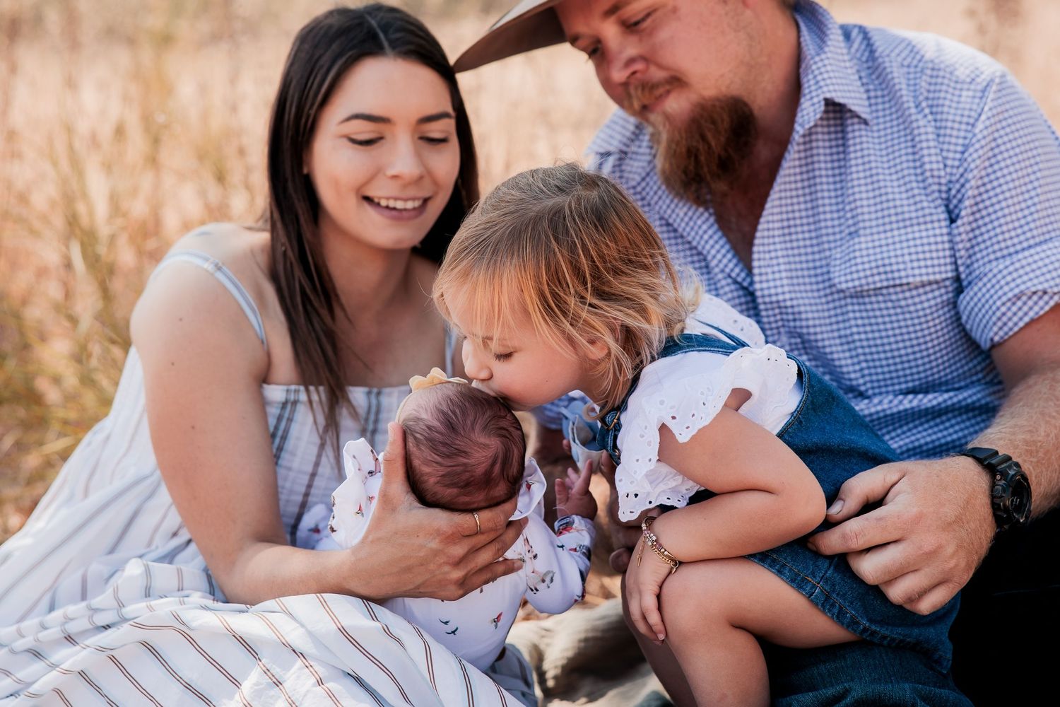 A family sits together in a field during golden hour as a toddler gently kisses a newborn baby.