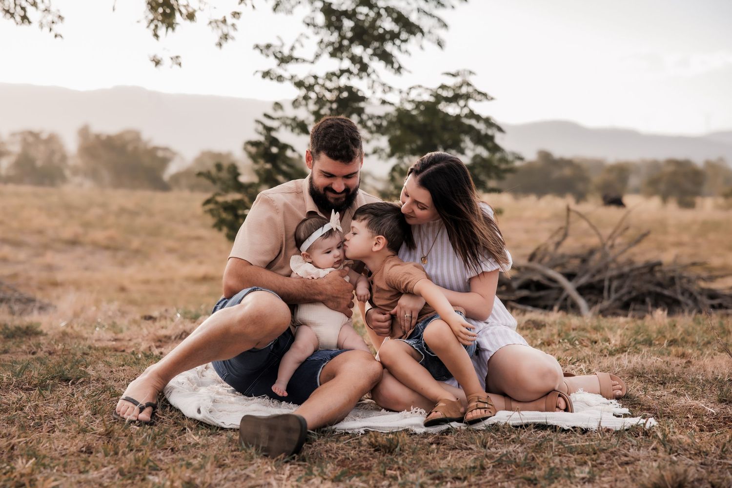 A family shares an intimate moment together while sitting on a blanket in a grassy field at sunset.