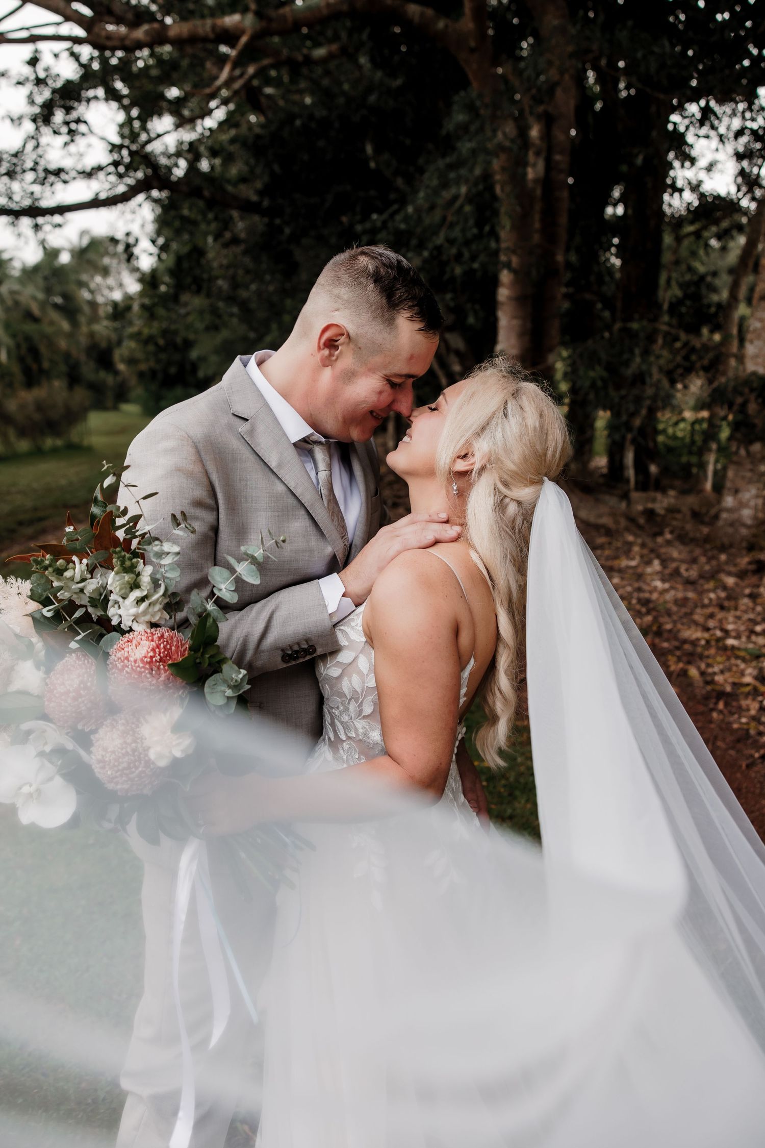 Bride in flowing white veil shares intimate moments with groom in outdoor woodland setting.