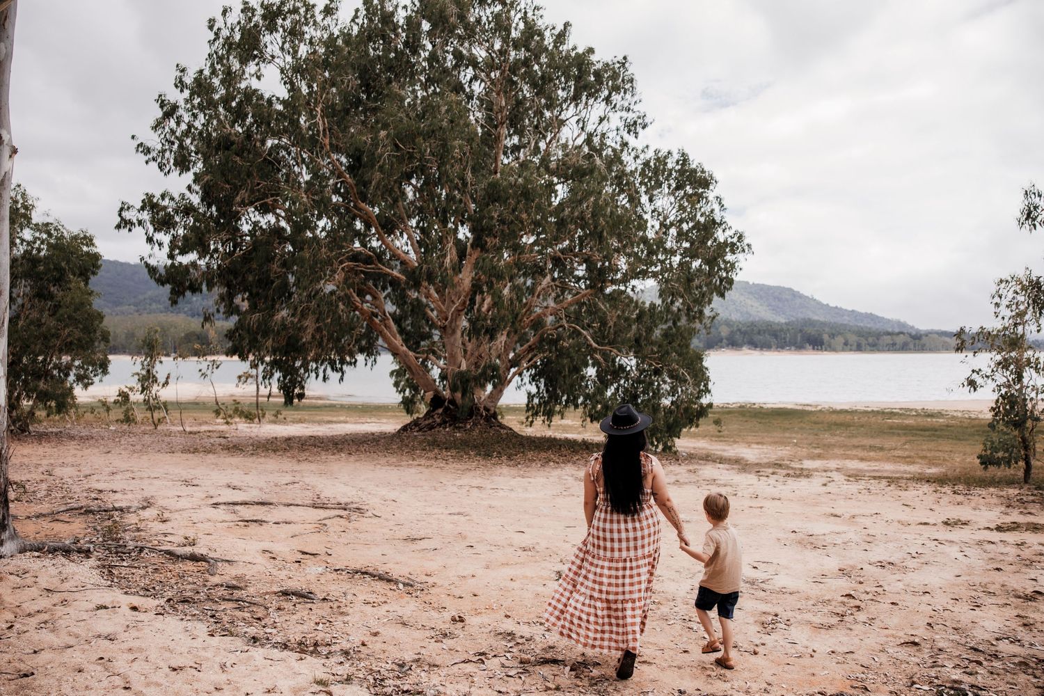 Parent and child walking together near a large tree by a lakeside on a cloudy day.