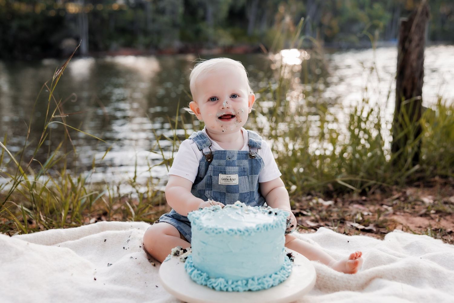 Baby in denim overalls smiling with blue birthday cake on white blanket by riverside.