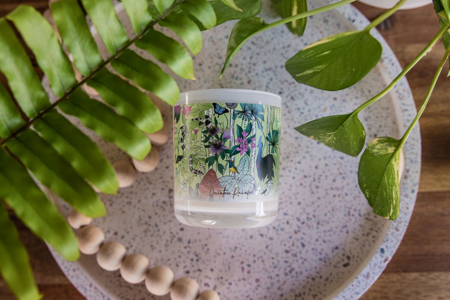 A decorative clear candle with floral designs sits on a gray speckled surface next to green fern leaves.