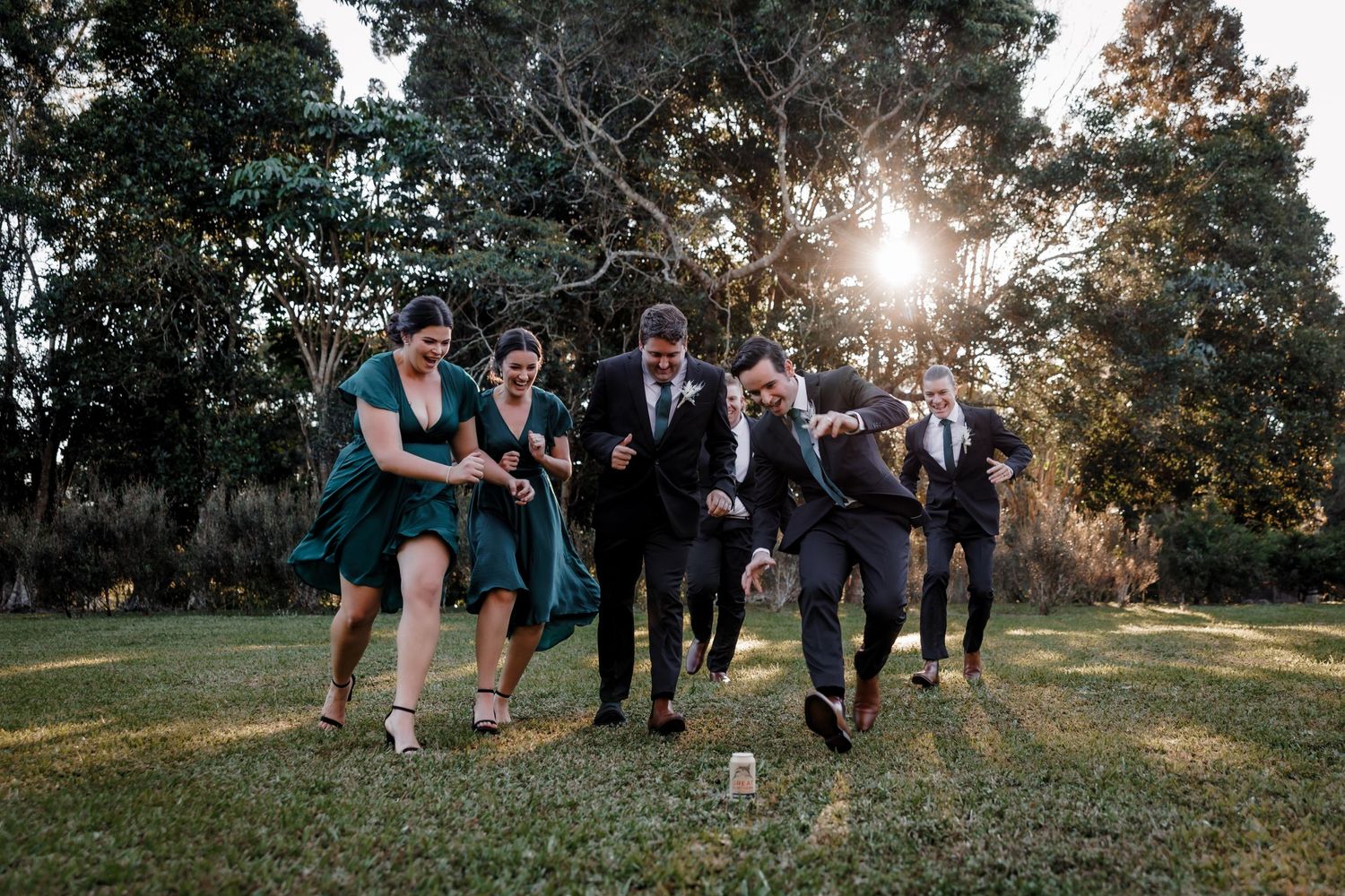 Wedding party members run and play together in a grassy field during sunset.