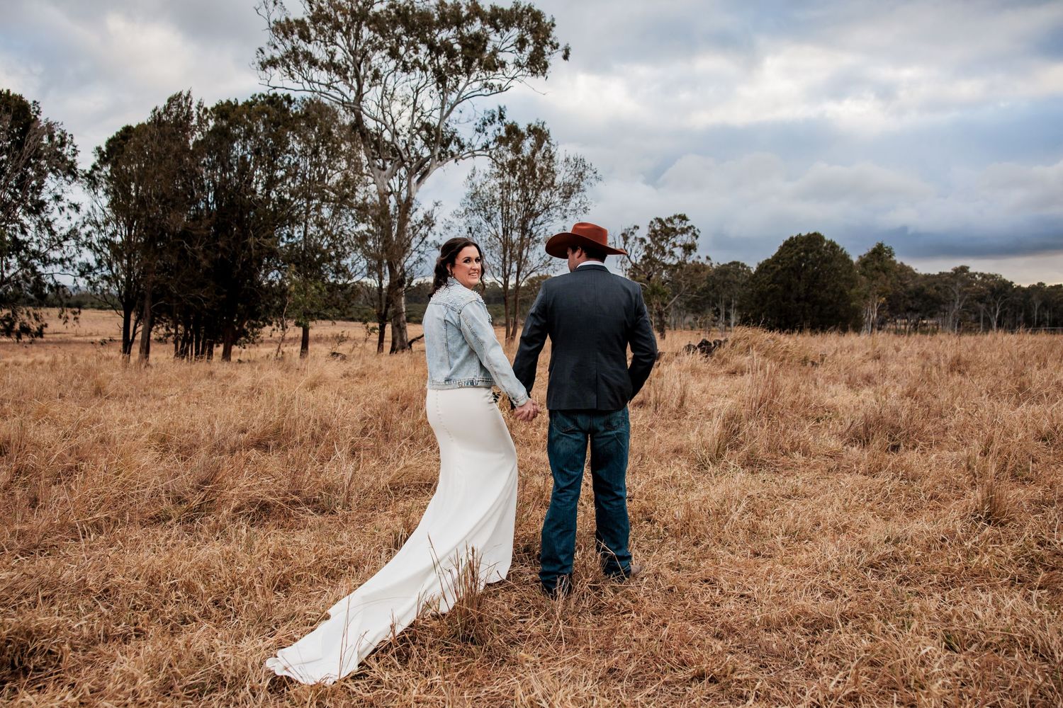 Couple in wedding attire walking through a golden field at sunset.
