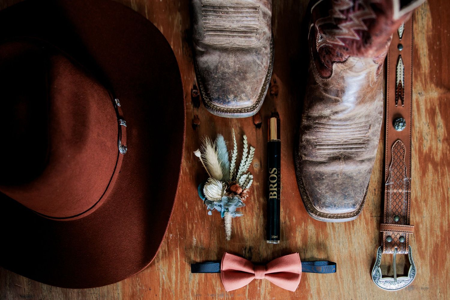 Wedding day details including boots, hat, bow tie and accessories arranged on wooden surface.
