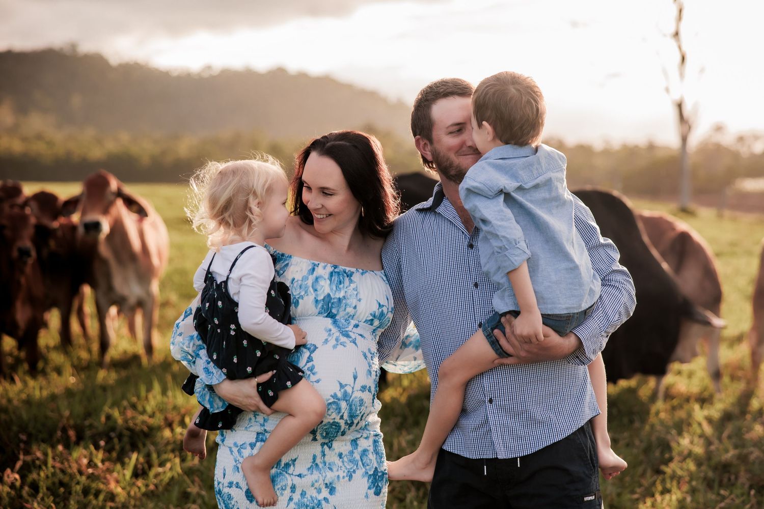 A family poses together in a field at sunset with horses grazing in the background.