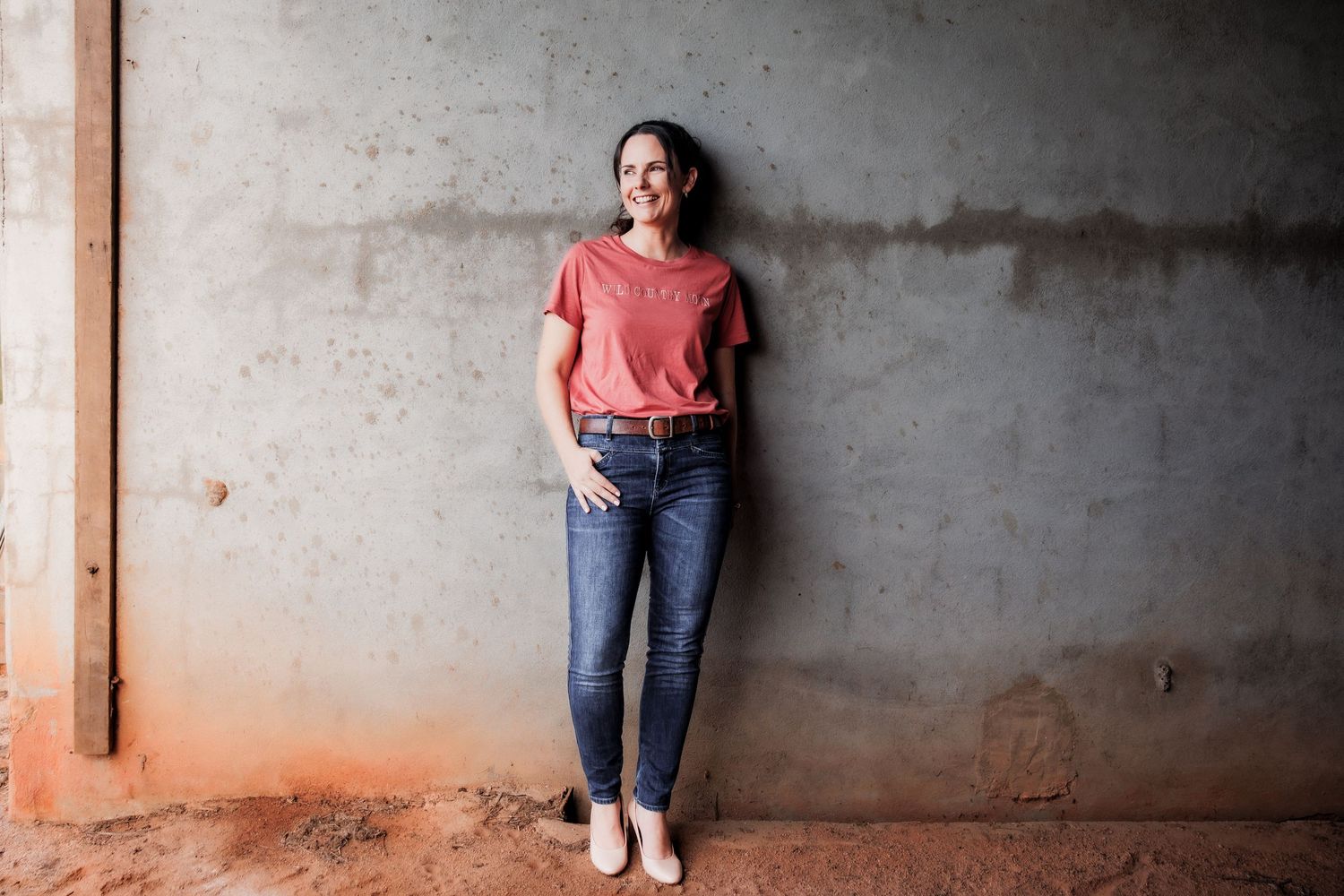A person in a coral top and jeans leans against a concrete wall in a casual, side-leaning pose.
