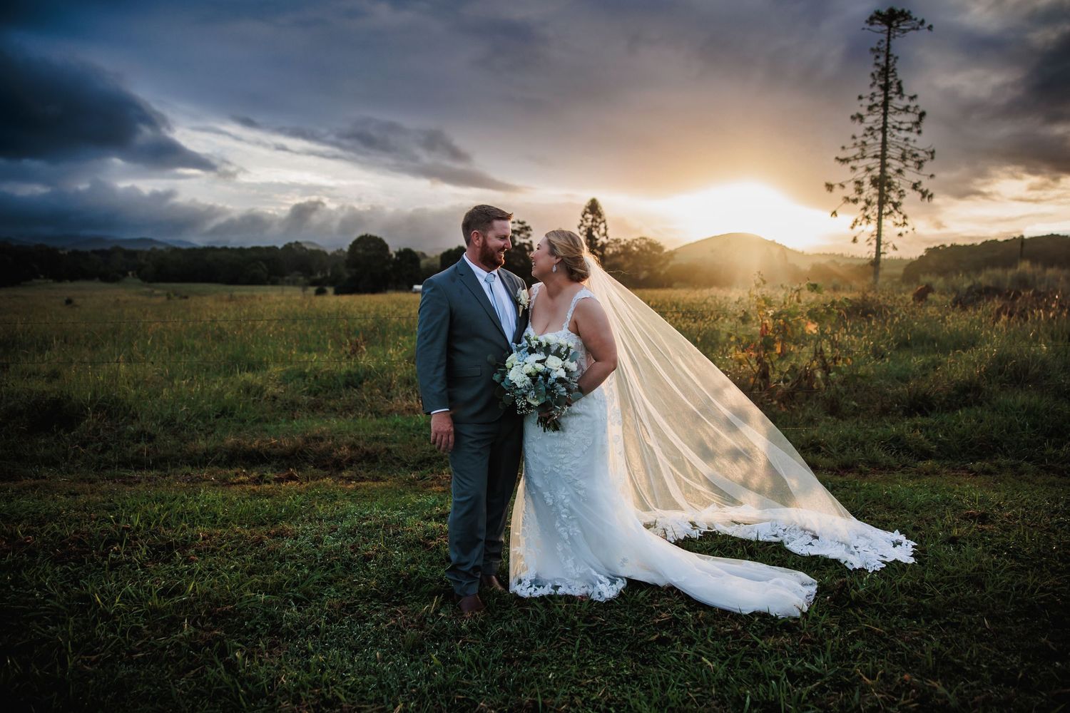 A wedding couple stands in a field at sunset with dramatic clouds and mountains in the background.