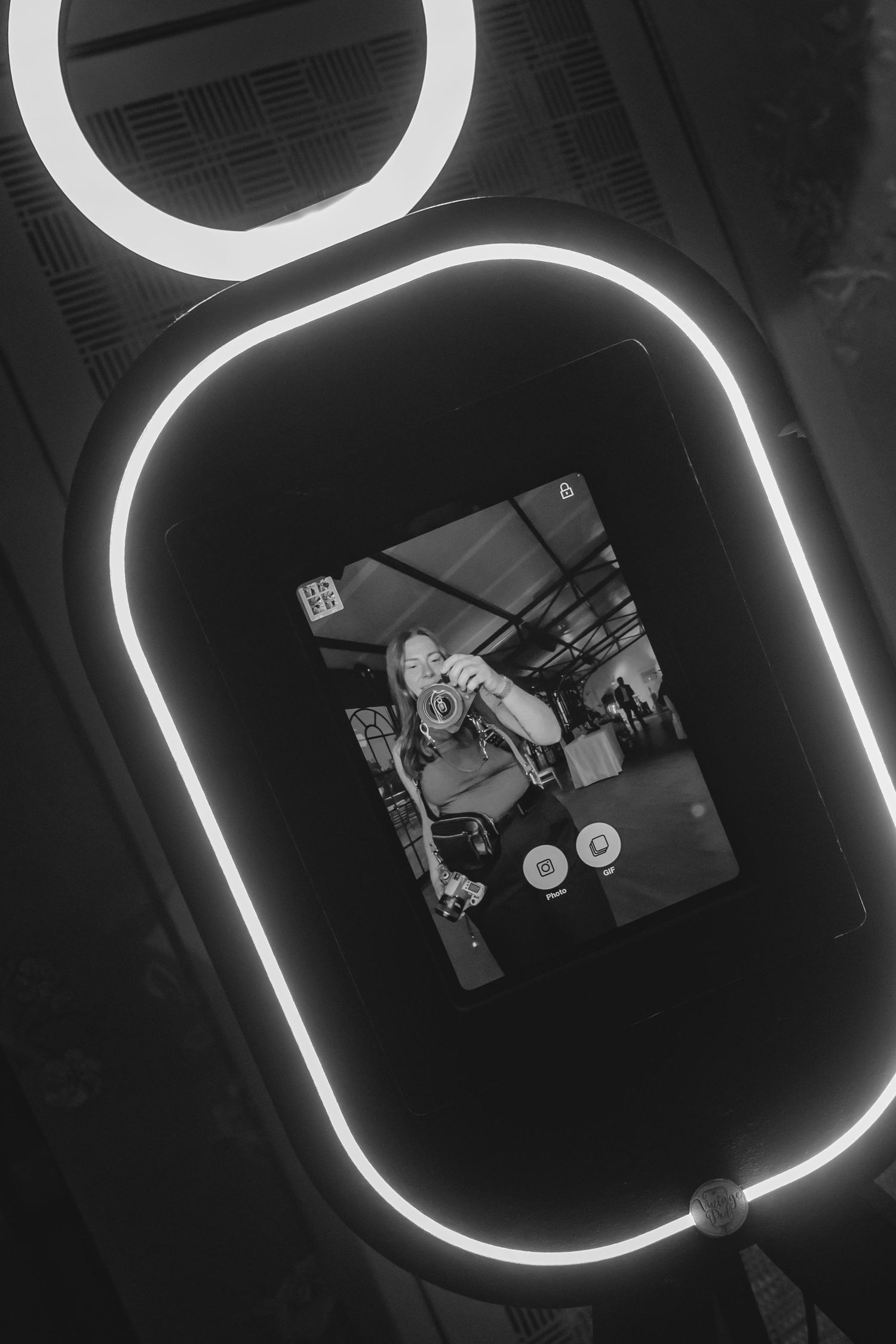 Black and white photo booth with glowing LED ring light frame displaying a camera screen selfie preview.