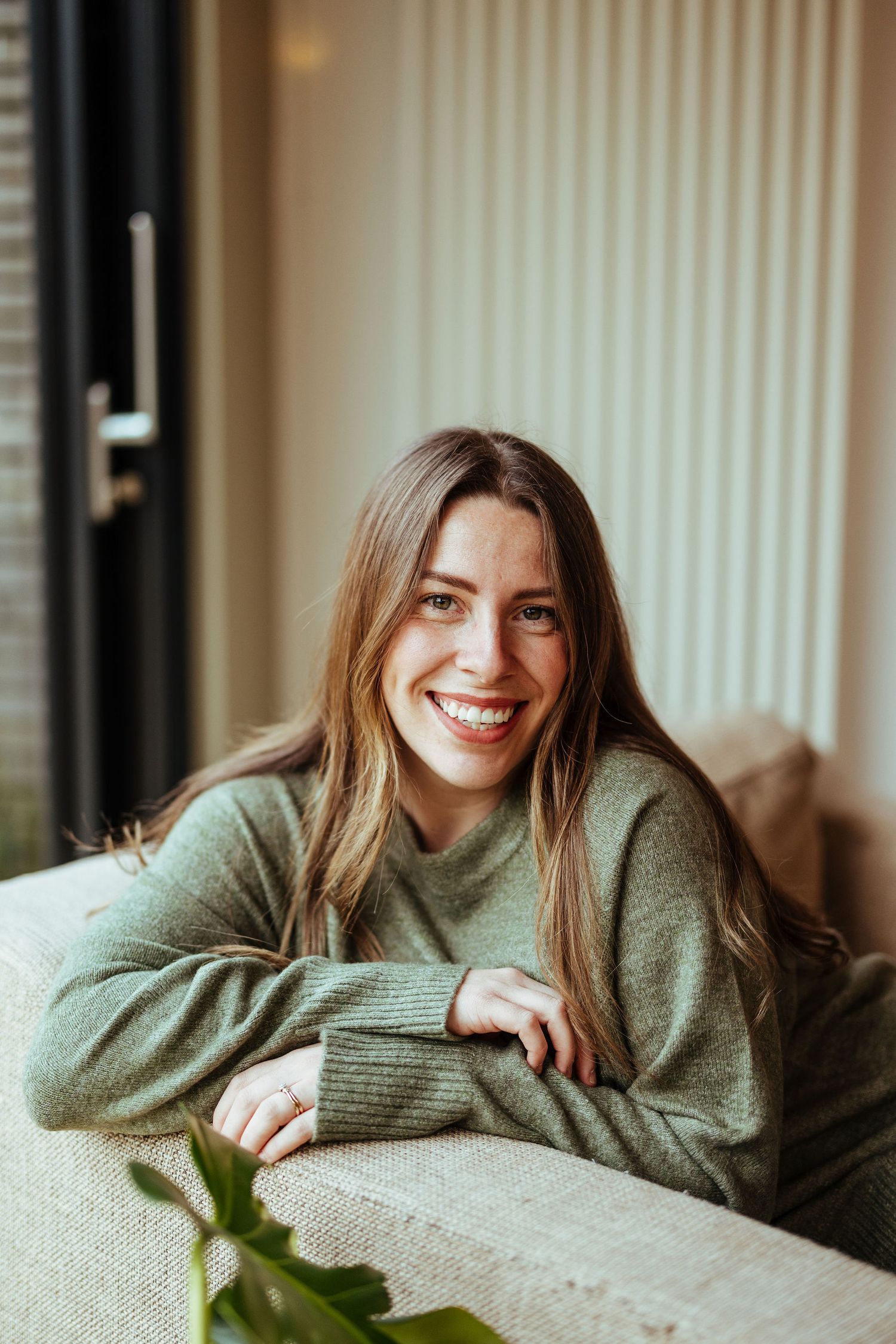 Smiling young woman with long brown hair wearing a green sweater, relaxing on a couch in a bright modern room.
