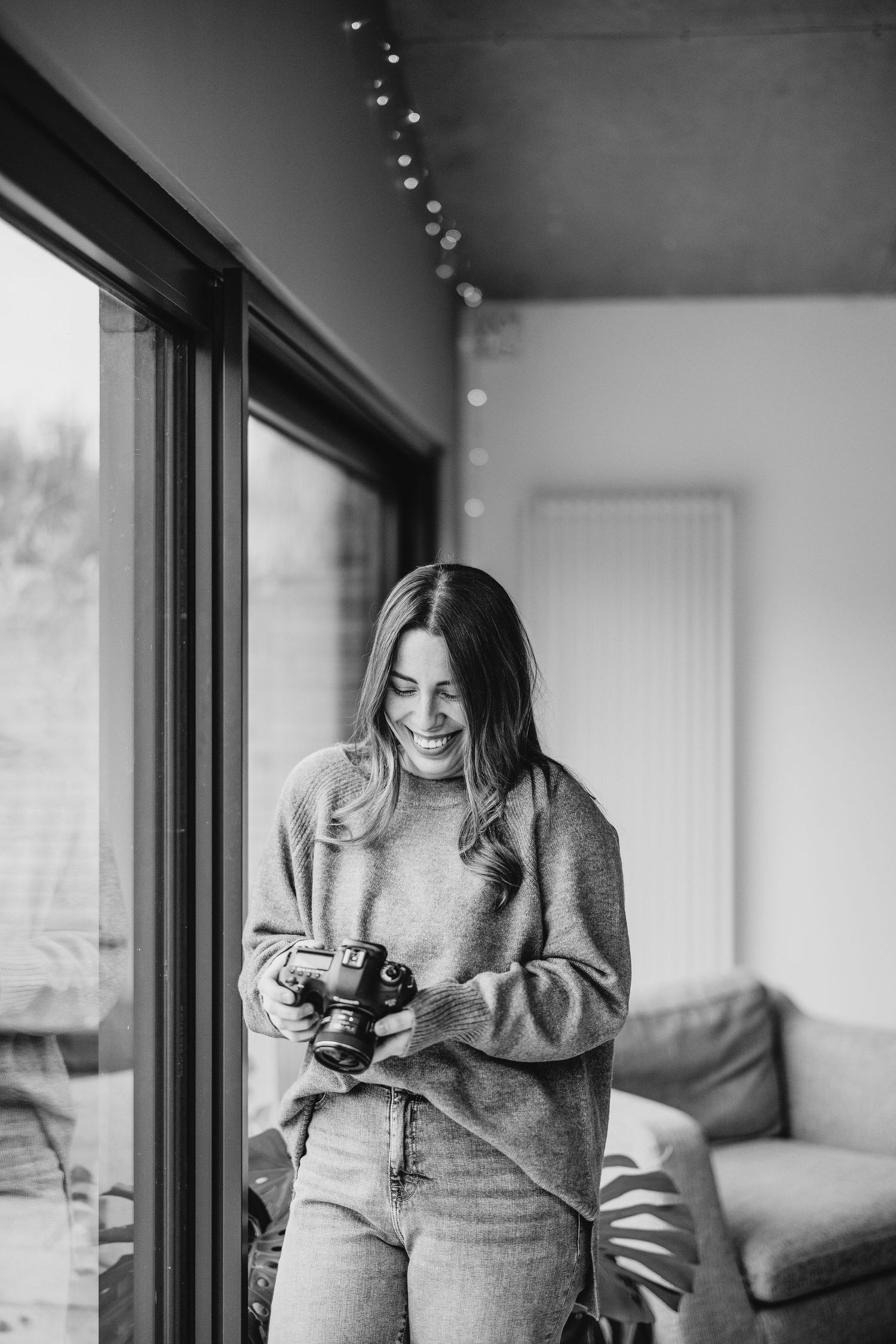 Black and white portrait of someone looking at a camera by a window with string lights.