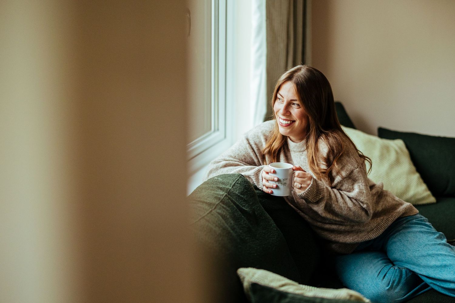 Woman smiling and holding a white mug while relaxing on a couch near a sunlit window at home.