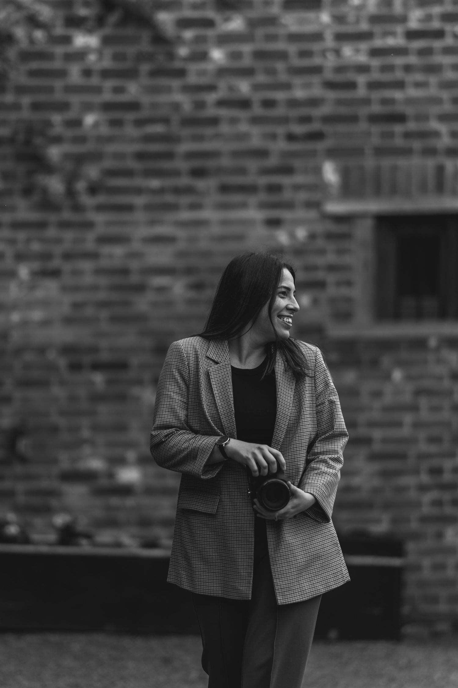 Black and white photo showing someone in a blazer holding a camera against a brick wall backdrop.