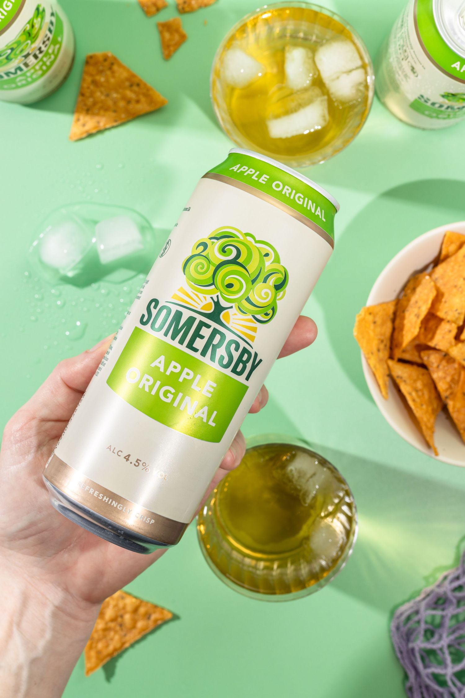 Hands holding green Somersby cider cans against mint background with snacks and ice cubes.