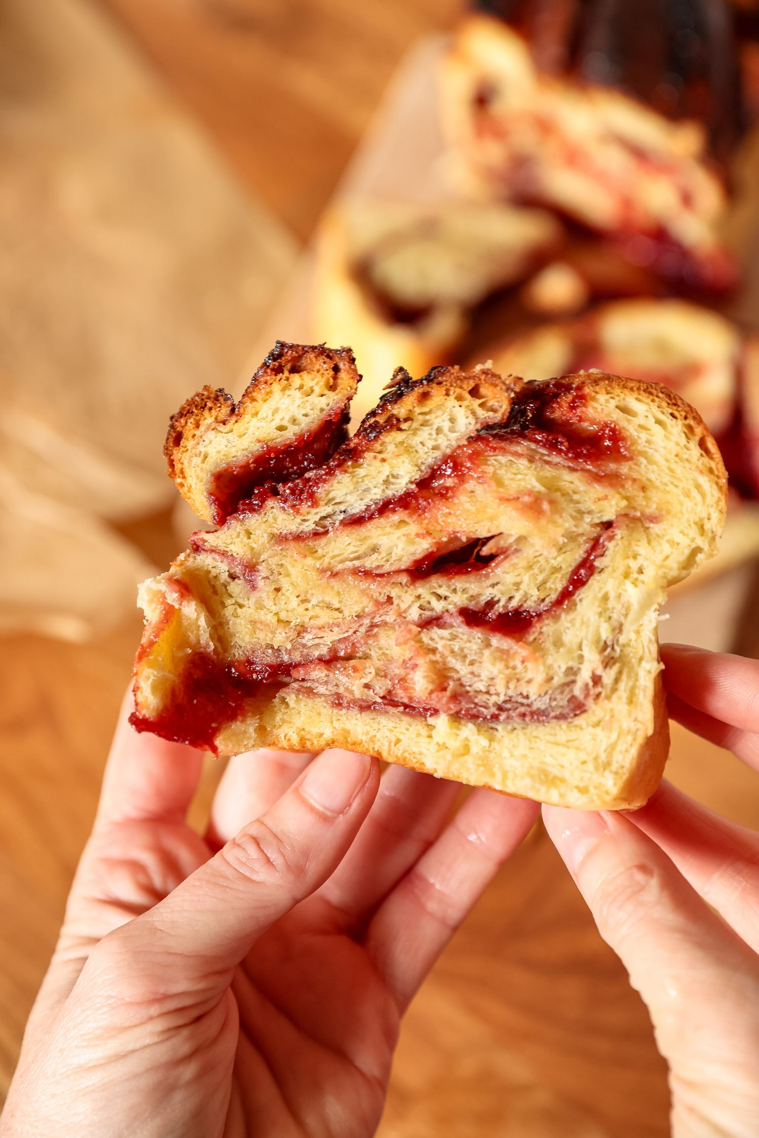 Hands holding a slice of freshly baked swirled babka bread with raspberry jam filling on a wooden table.