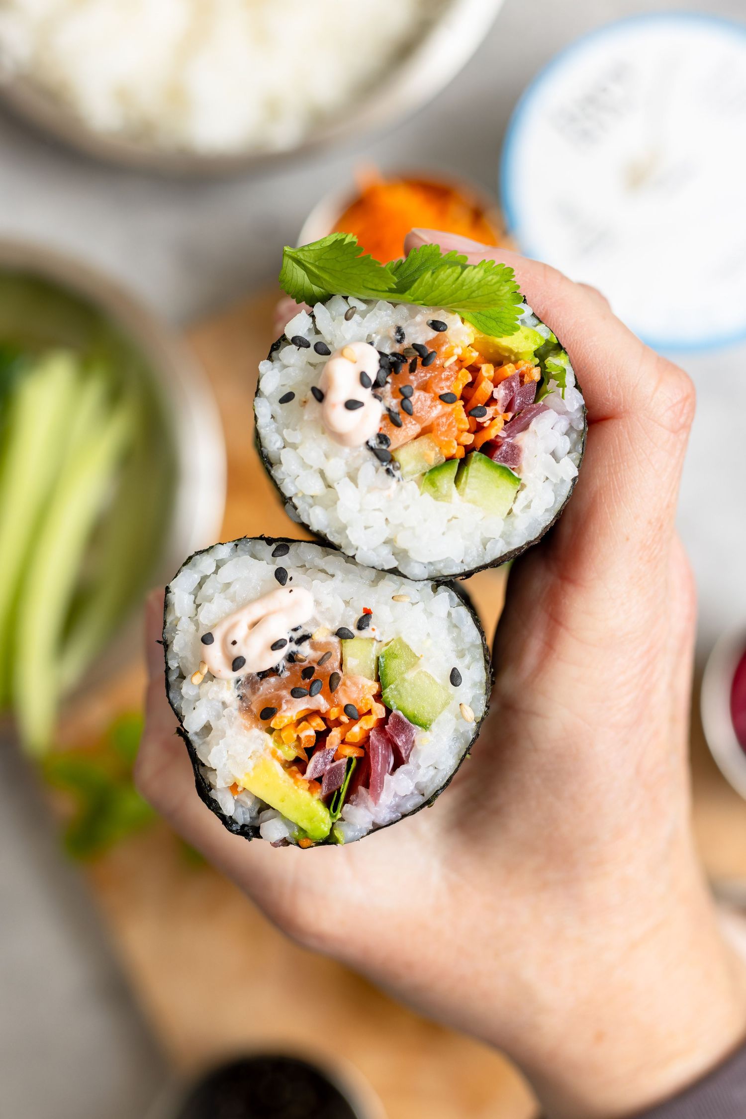 Hand holding a sushi burrito cut in half, filled with rice, avocado, vegetables, and spicy sauce, wrapped in nori seaweed.