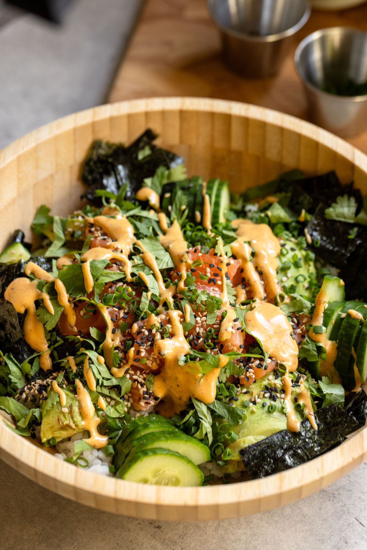 Wooden bowl filled with a poke bowl topped with spicy mayo, seaweed, cucumber, greens, and sesame seeds.