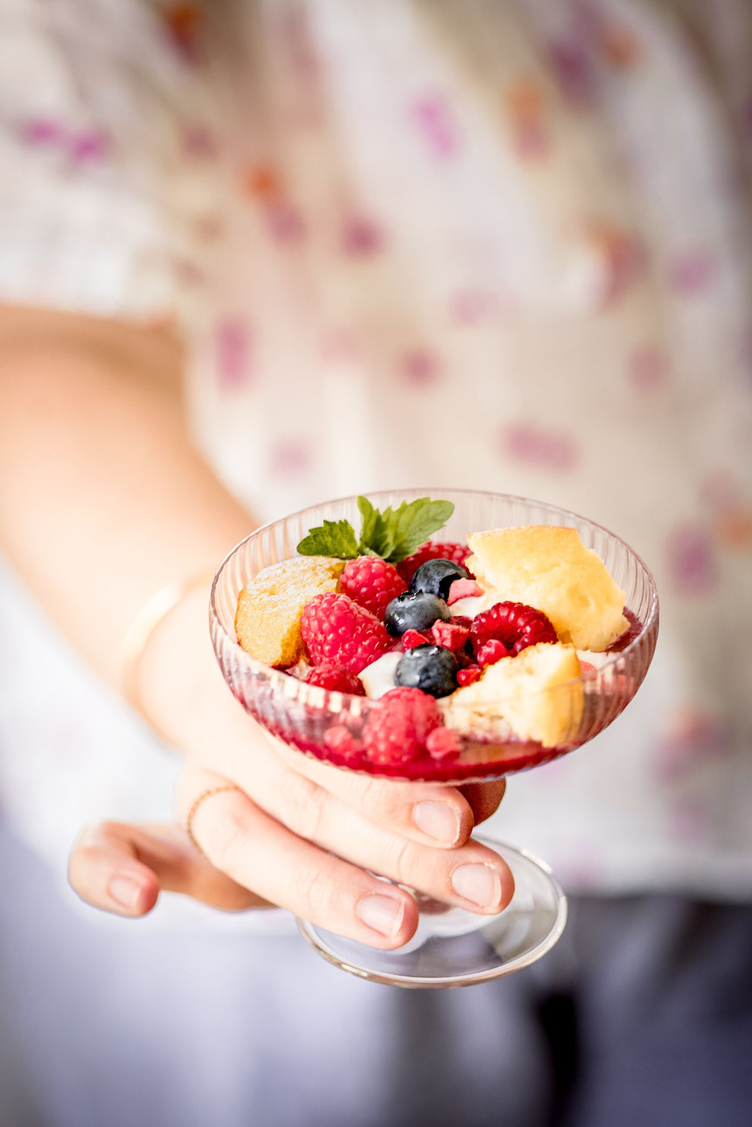Hand holding a glass dessert cup filled with fresh raspberries, blueberries, and sponge cake pieces garnished with mint.