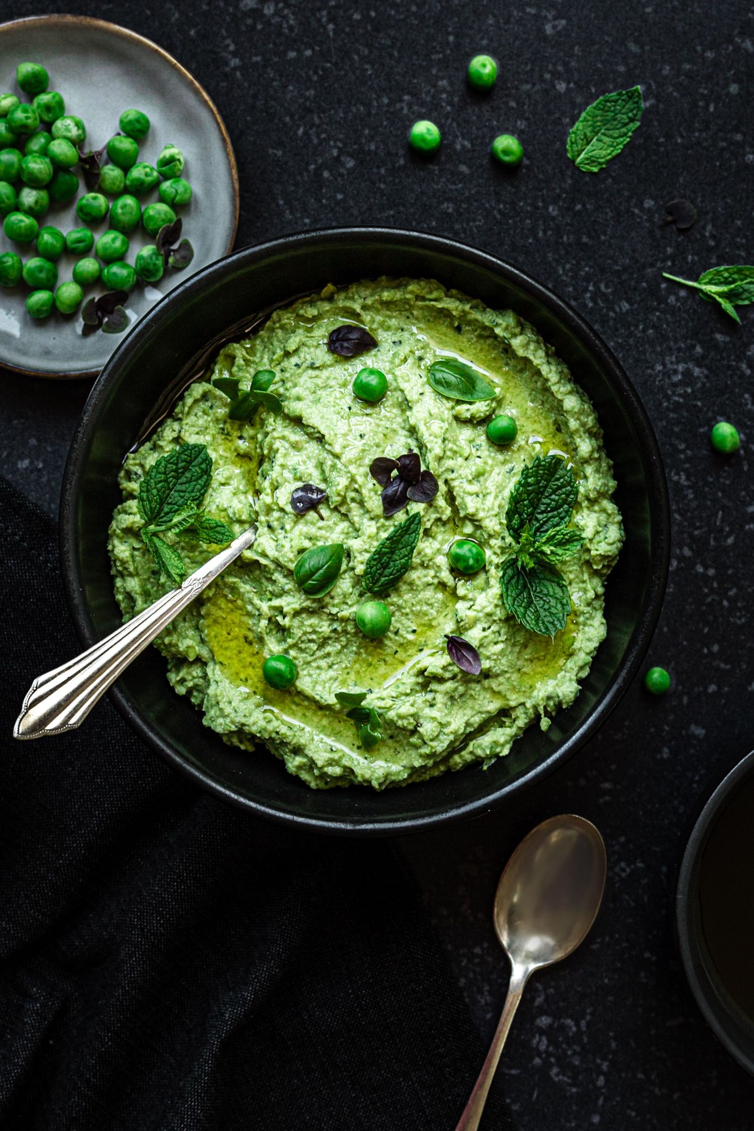 Green pea hummus in a black bowl topped with mint leaves, olive oil, and fresh peas on a dark background.