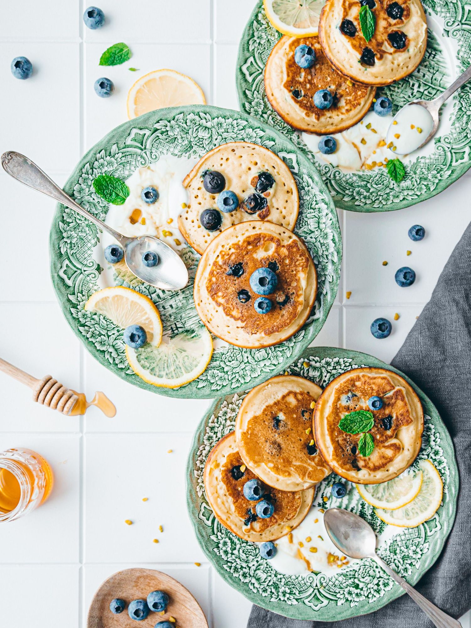 Three green plates of fluffy pancakes topped with blueberries, lemon slices, mint, and honey on a white tile surface.