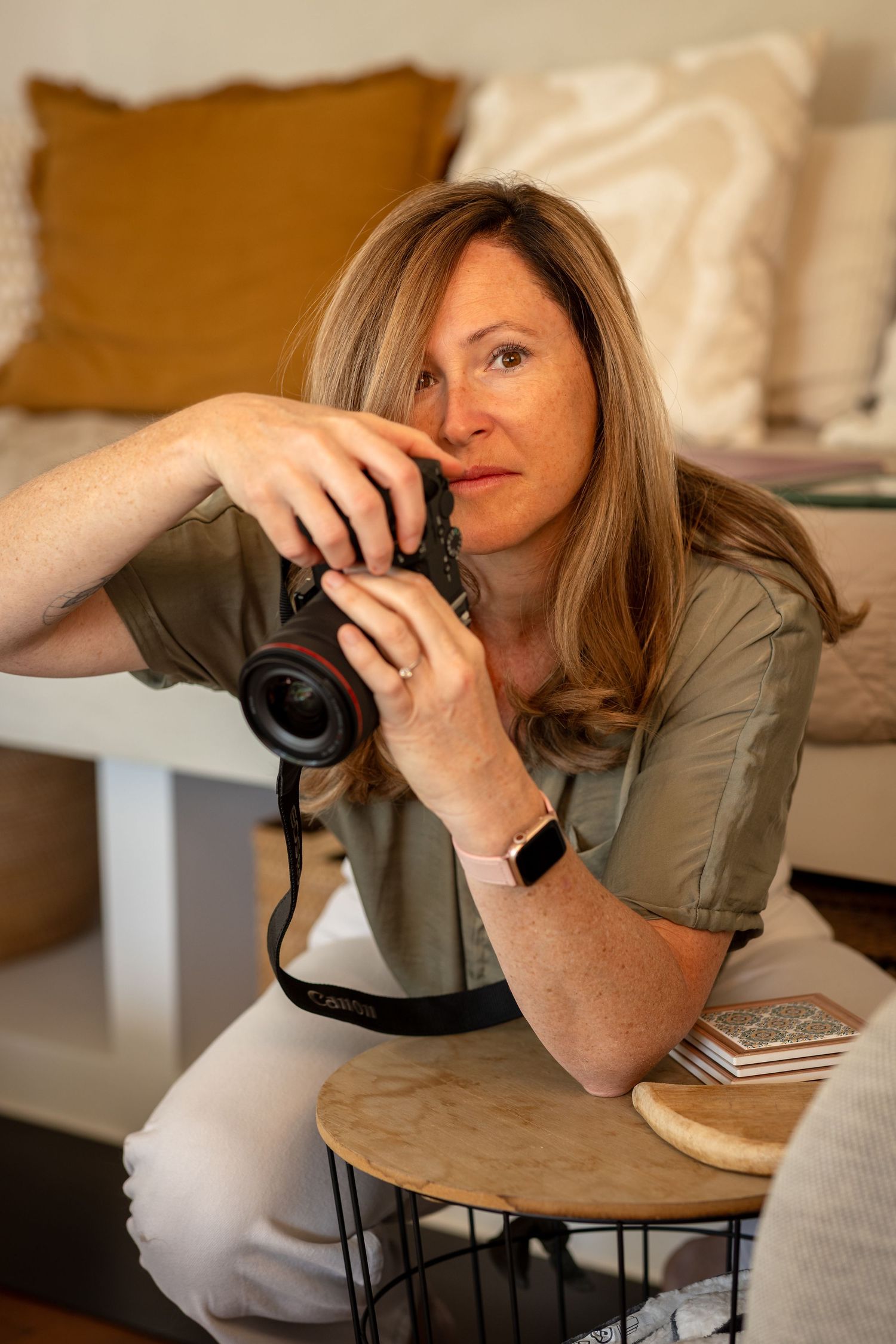 Woman holding a Canon camera up to her eye while sitting in a cozy living room, wearing a green shirt and smartwatch.