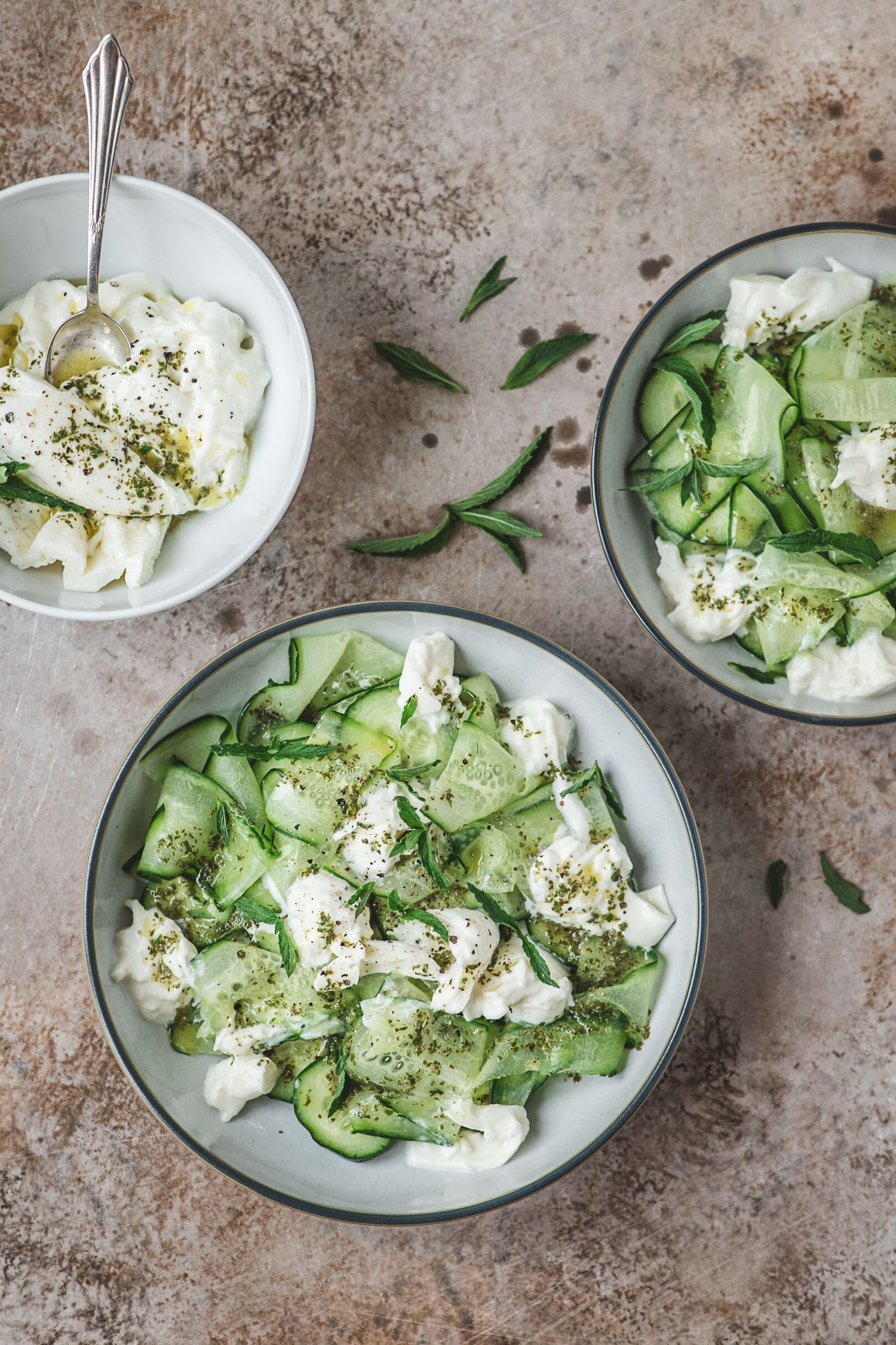 Three bowls of fresh cucumber salad with creamy burrata cheese, herbs, and seasoning on a rustic stone surface.