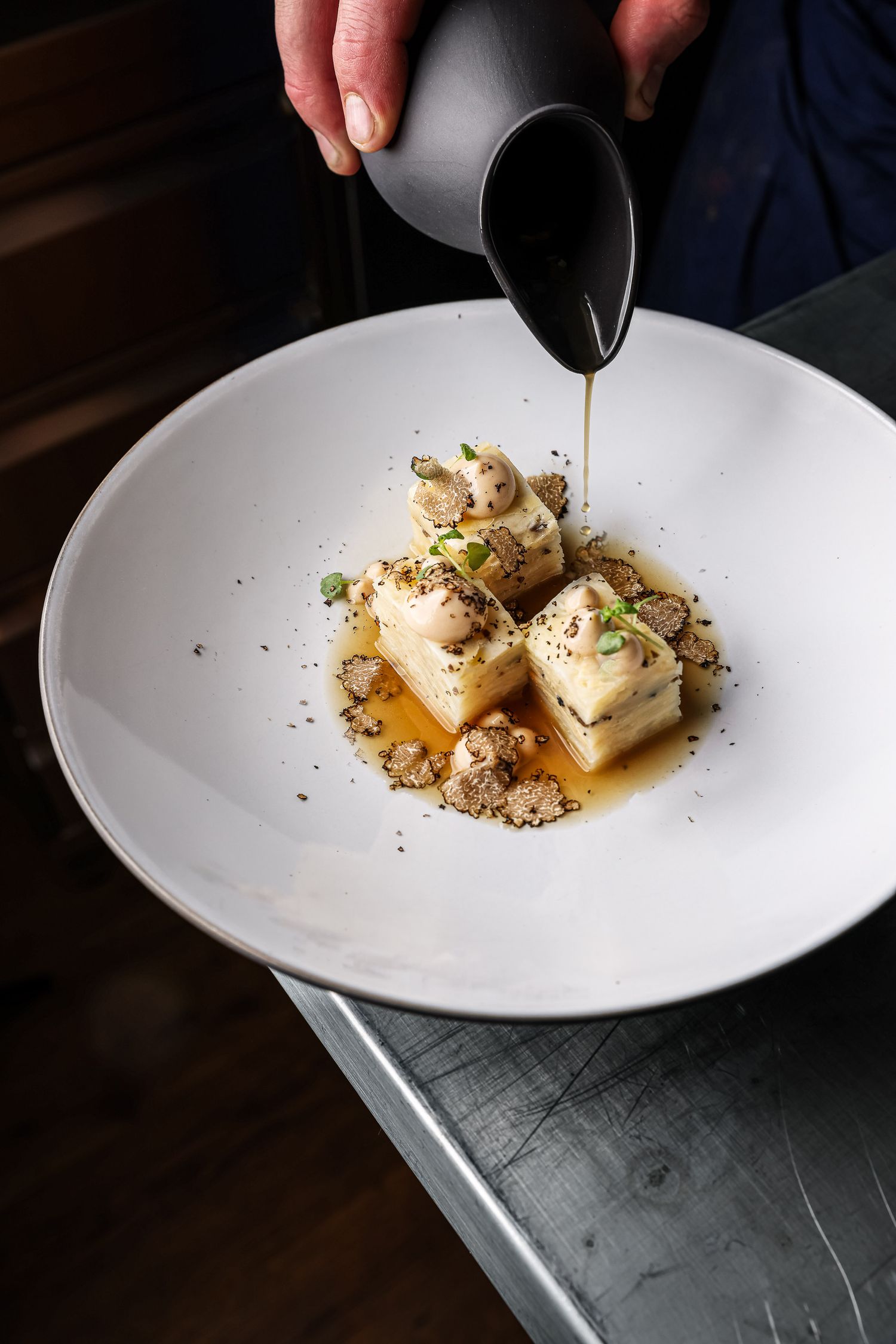 Chef pouring golden broth over elegant tofu cubes garnished with mushrooms and herbs on a white plate.