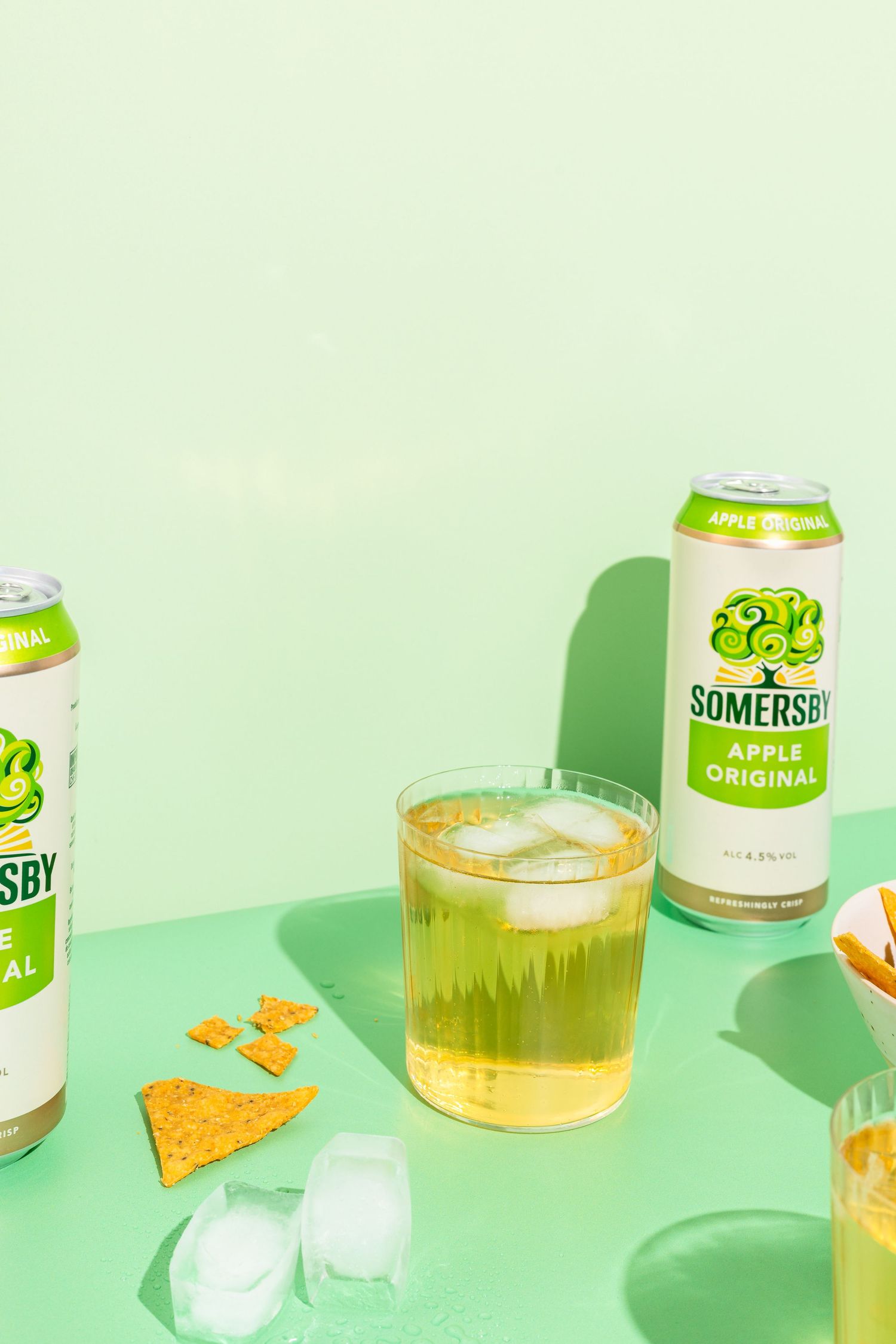 Cans of Somersby cider and glasses of beer arranged on mint green background with scattered ice cubes and snacks.