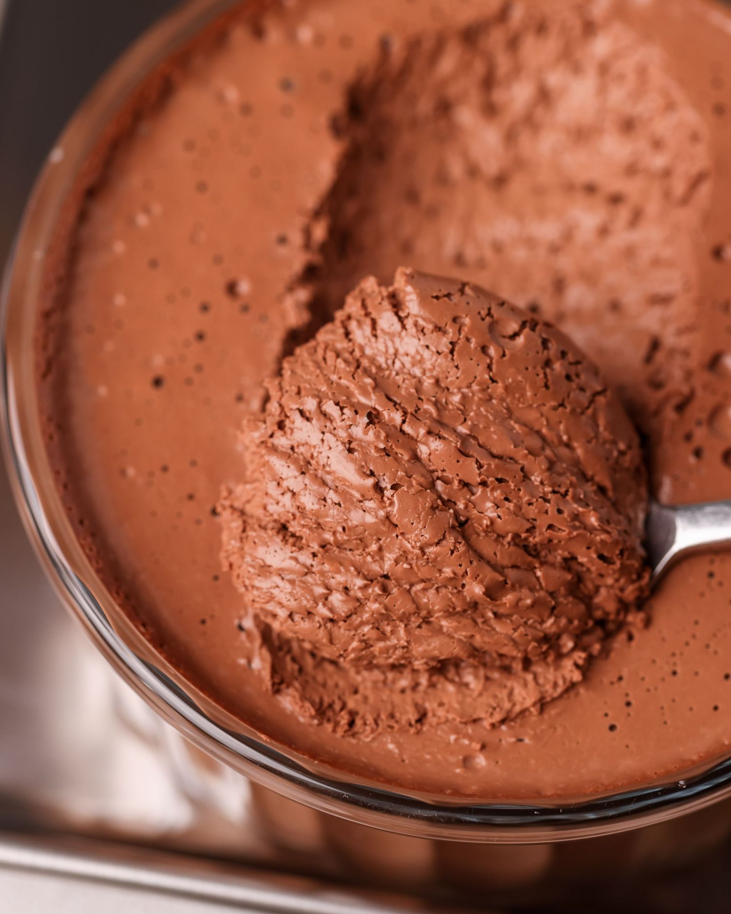 A spoon scooping rich chocolate mousse or pudding from a glass bowl showing its smooth creamy texture.