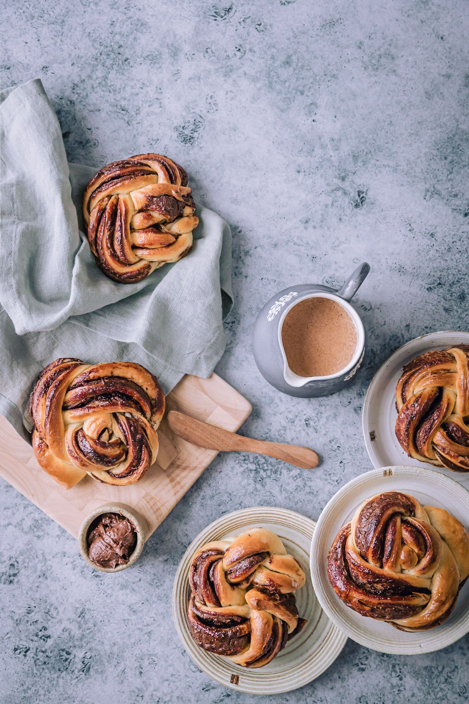 Freshly baked chocolate swirl buns on plates and wooden board with a small pitcher of sauce on gray surface.