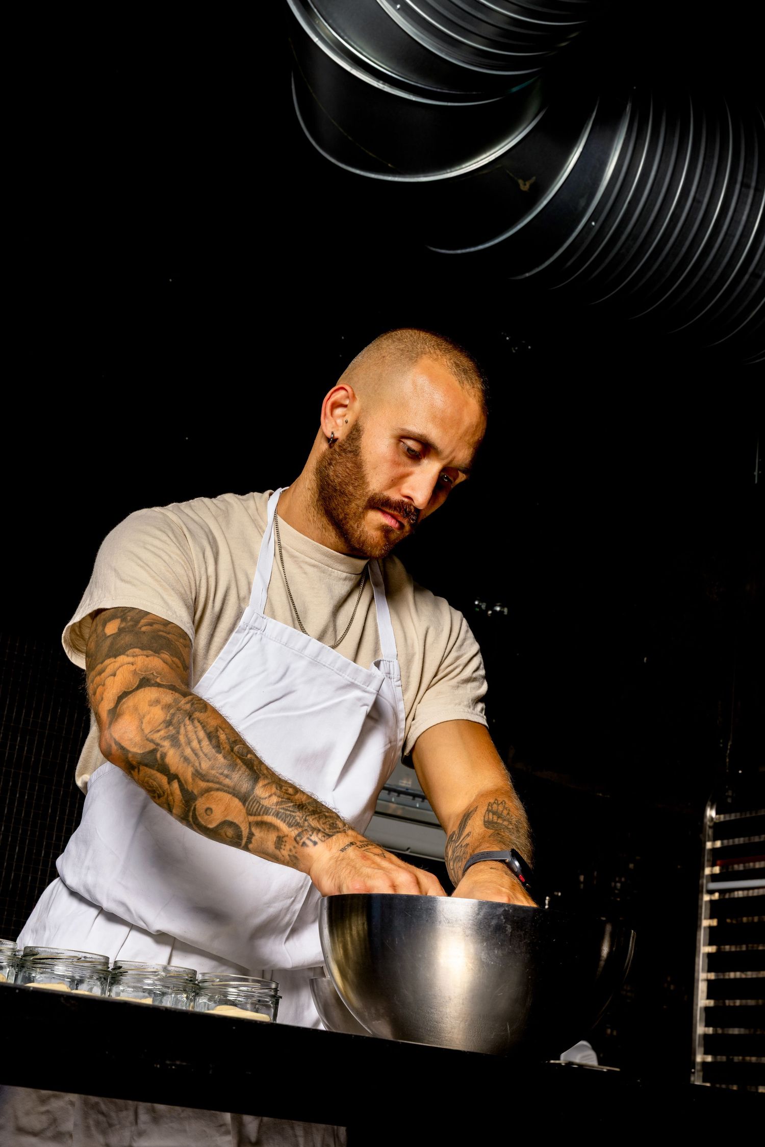 Tattooed male chef in white apron mixing ingredients in a stainless steel bowl in a professional kitchen.
