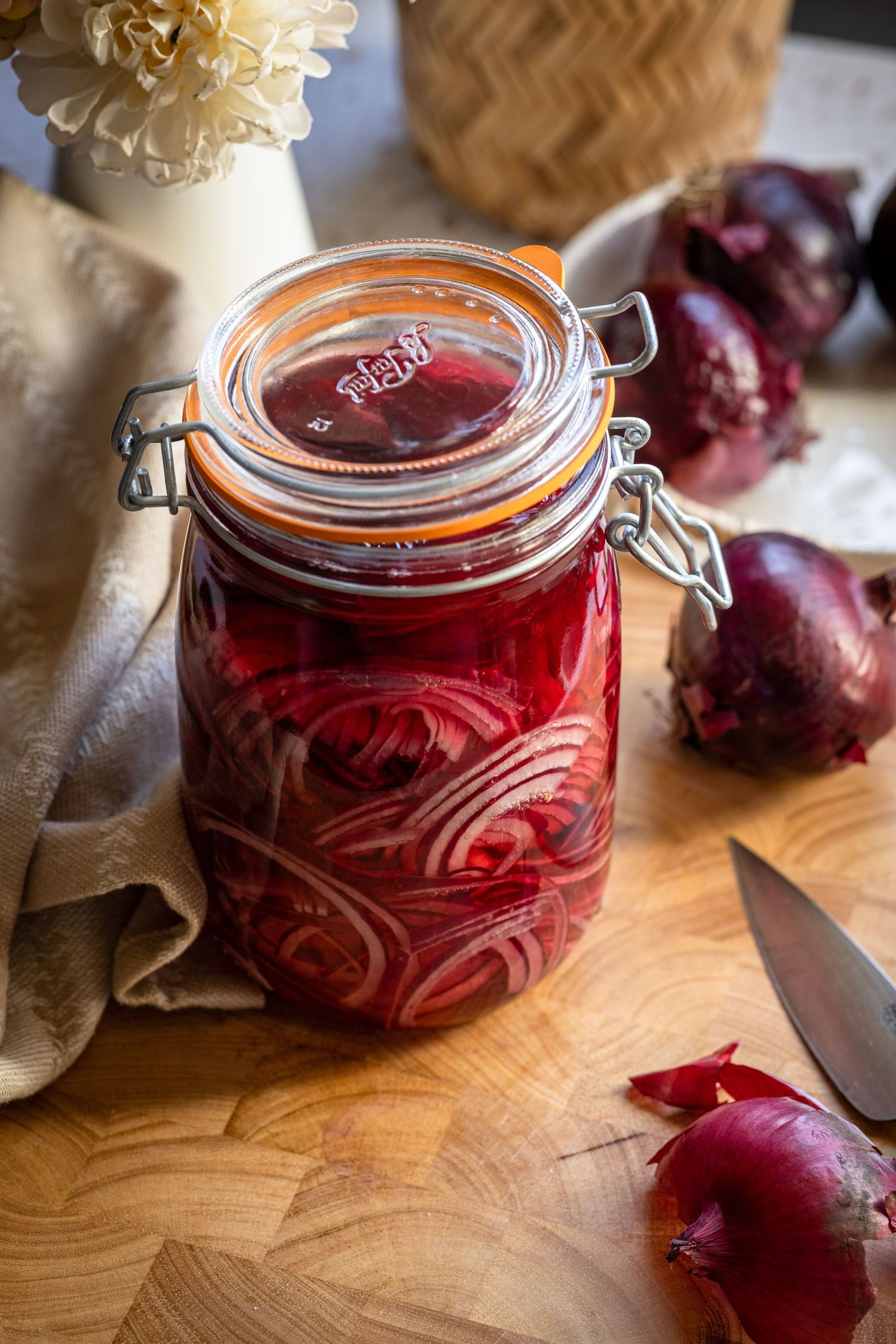 Glass jar of vibrant red pickled onions on a wooden cutting board with fresh red onions nearby.