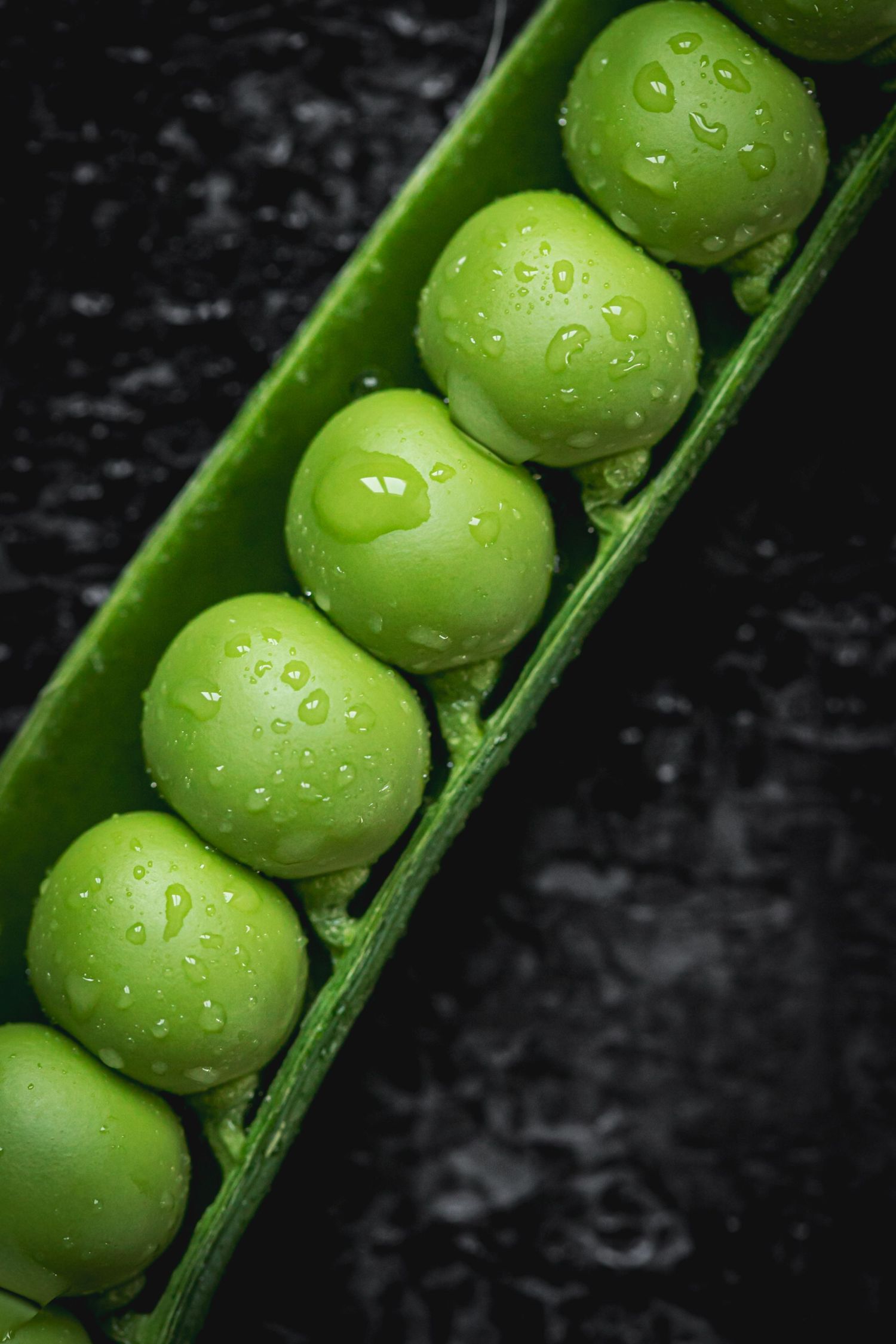 Close-up of fresh green peas in an open pod with water droplets on dark background.
